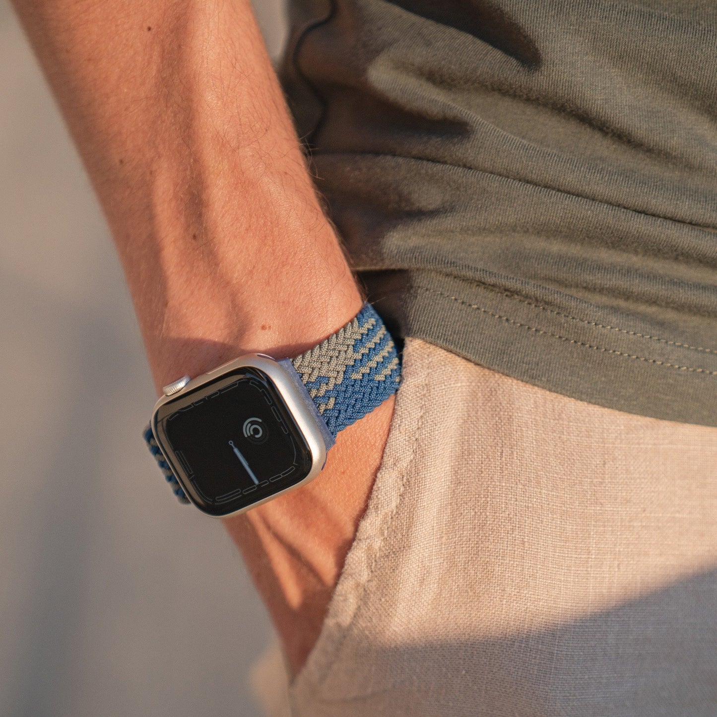 Eine Person in einem grauen Hemd und einer beigen Hose hat ihre linke Hand in der Tasche und zeigt ein geflochtenes Flex Loop TwoTone Armband der Apple Watch an ihrem Handgelenk.