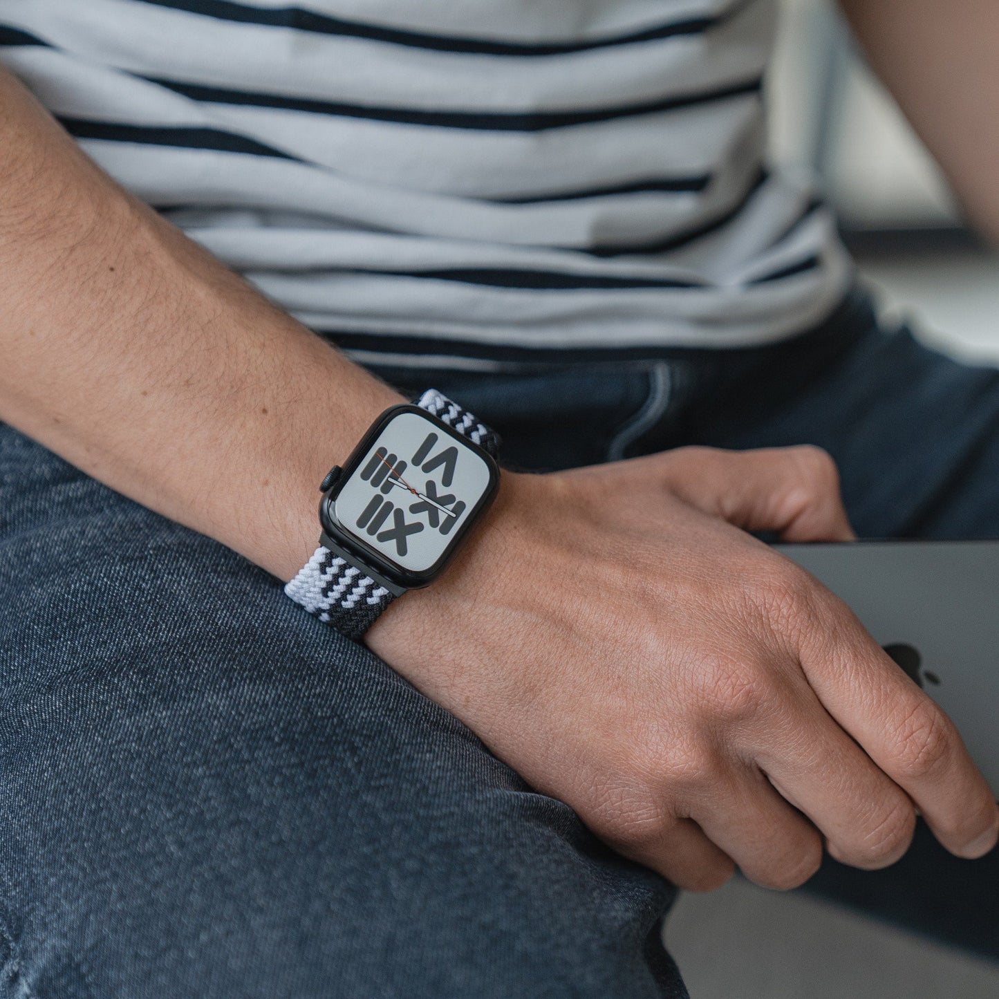 Eine Person trägt ein geflochtenes Flex Loop TwoTone-Armband für die Apple Watch mit Edelstahlverschluss und einem Zifferblatt mit römischen Ziffern und hält einen geschlossenen Laptop oder ein Tablet in der Hand. Sie ist mit Blue Jeans und einem gestreiften Hemd bekleidet.