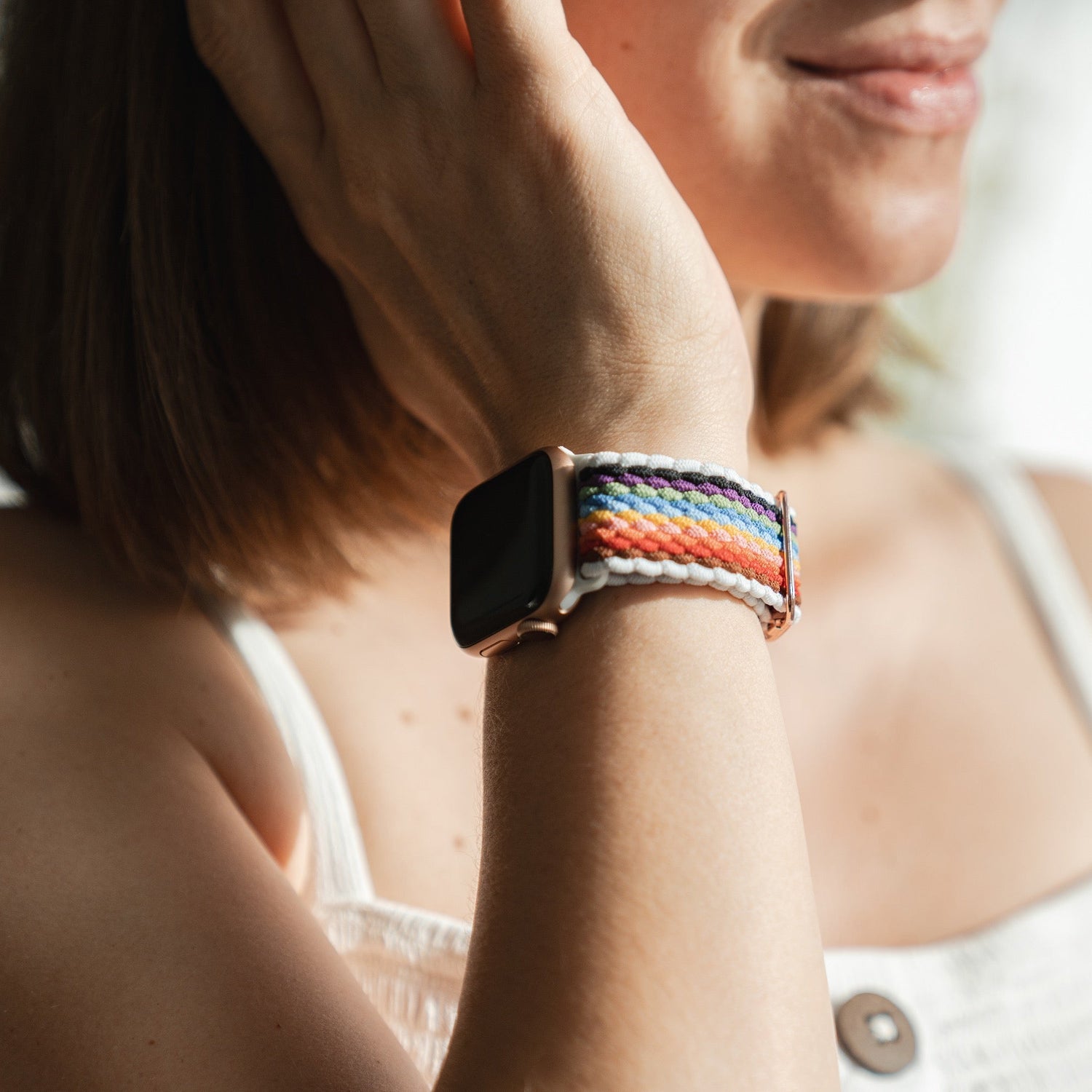 Eine Person in einem ärmellosen weißen Oberteil präsentiert ihr Handgelenk mit dem geflochtenen Flex Loop Pride Armband von arktisband, einem gewebten regenbogenfarbenen Armband für die Apple Watch. Sie lächelt leicht und berührt ihr Gesicht.