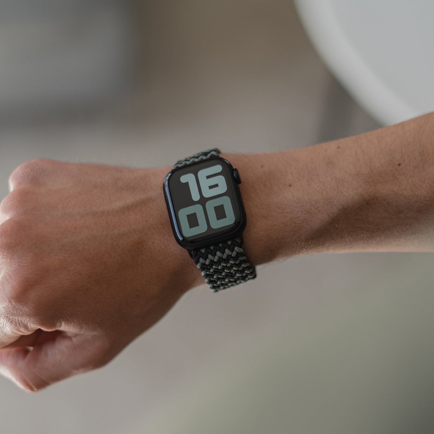 Am linken Handgelenk trägt eine Person ein geflochtenes Flex Loop TwoTone Armband in Schwarz und Grau. Die Apple Watch zeigt 16:00 Uhr in großen weißen Ziffern auf einem schwarzen Bildschirm an.