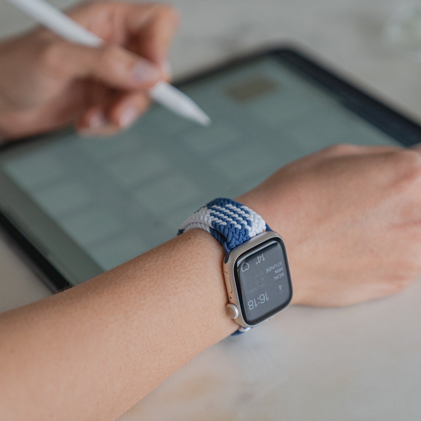 Eine Person, die ein Arktisband Apple Watch geflochtenes Flex Loop TwoTone Armband trägt, benutzt einen Stift auf einem Tablet. Das Arktisband zeigt Uhrzeit und Benachrichtigungen an, während im Hintergrund der unscharfe Tablet-Bildschirm sichtbar ist.