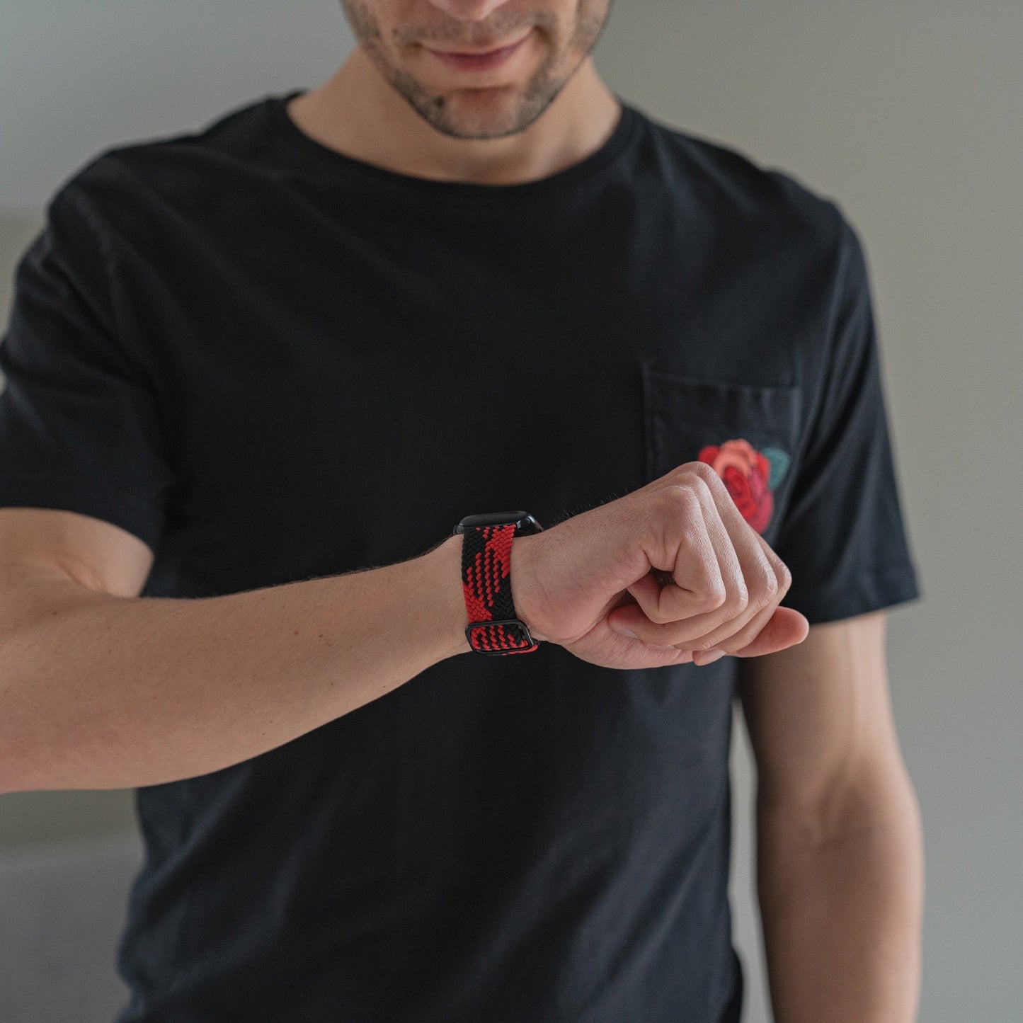 Eine Person in einem schwarzen T-Shirt mit einer roten Rosentasche blickt auf ihr Handgelenk und trägt das geflochtene Flex Loop TwoTone Armband von arktisband für die Apple Watch mit rot-schwarzem Muster und Edelstahlverschluss.