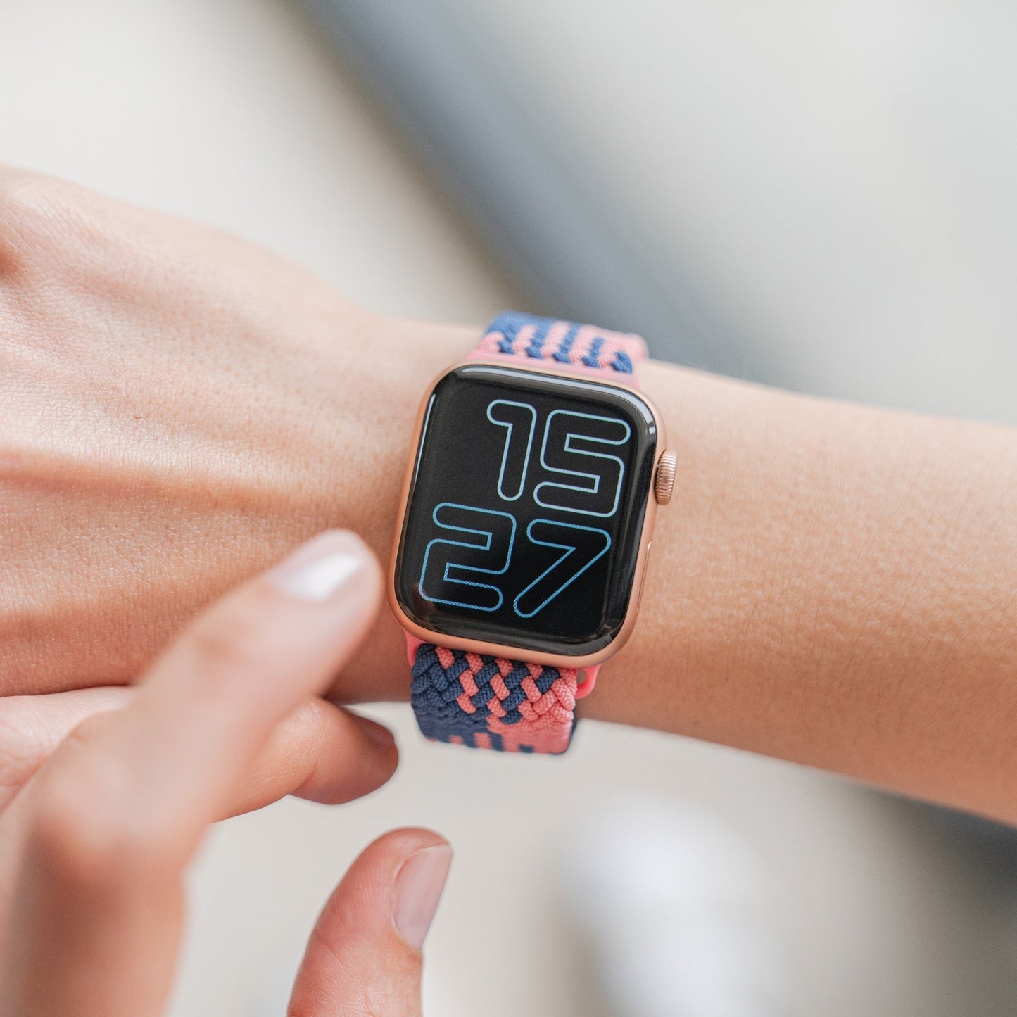 Eine Person trägt ein geflochtenes Flex Loop TwoTone Armband mit Arktisband für die Apple Watch mit einer großen Digitalanzeige, die 15:27 anzeigt, und einem Edelstahlverschluss, während eine andere Hand nach dem Bildschirm greift.