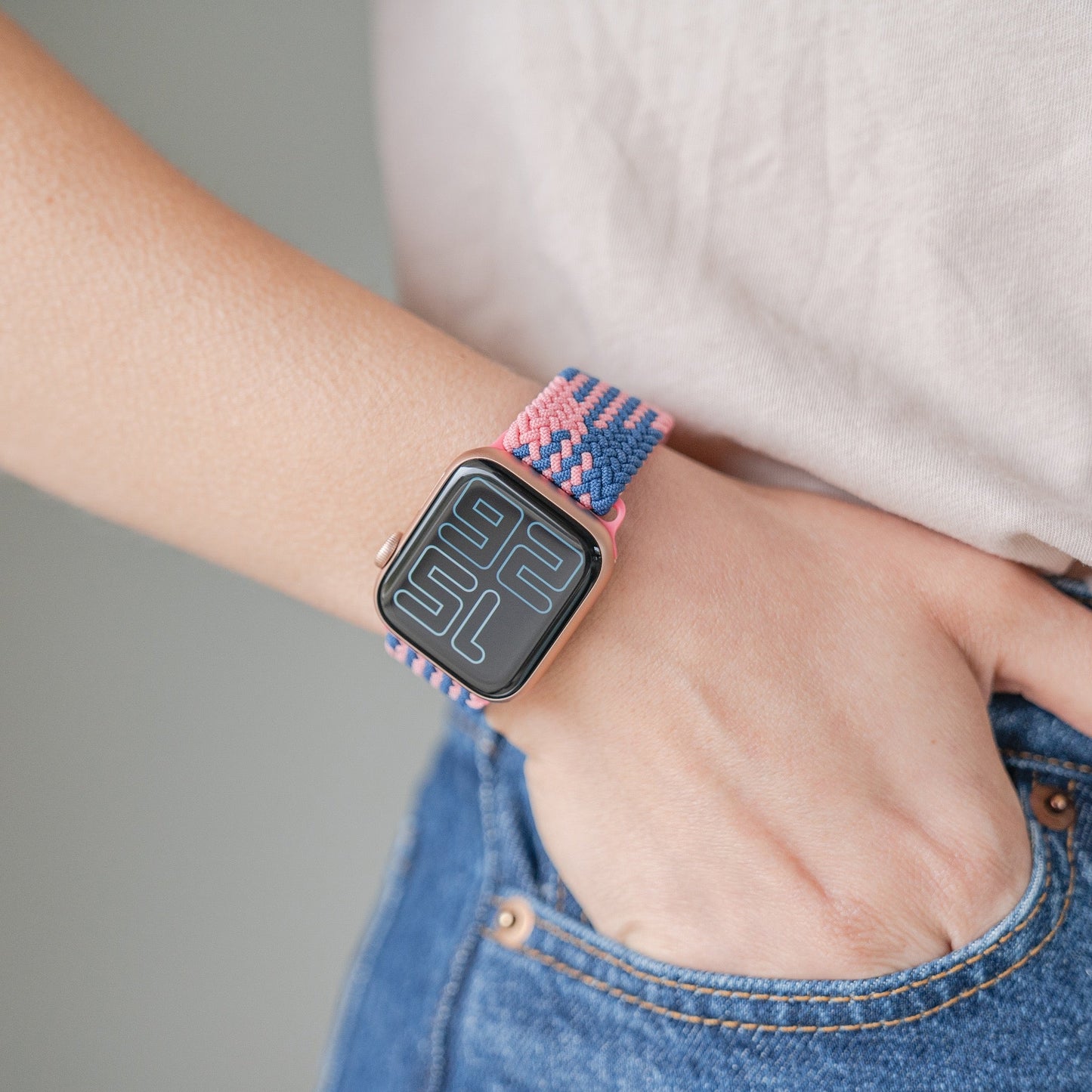 Jemand trägt Bluejeans, ein helles Hemd und ein geflochtenes Flex Loop TwoTone Armband von Arktisband für die Apple Watch in Pink und Blau; die Hand steckt in der Tasche und die Uhr zeigt ein minimalistisches digitales Zifferblatt.