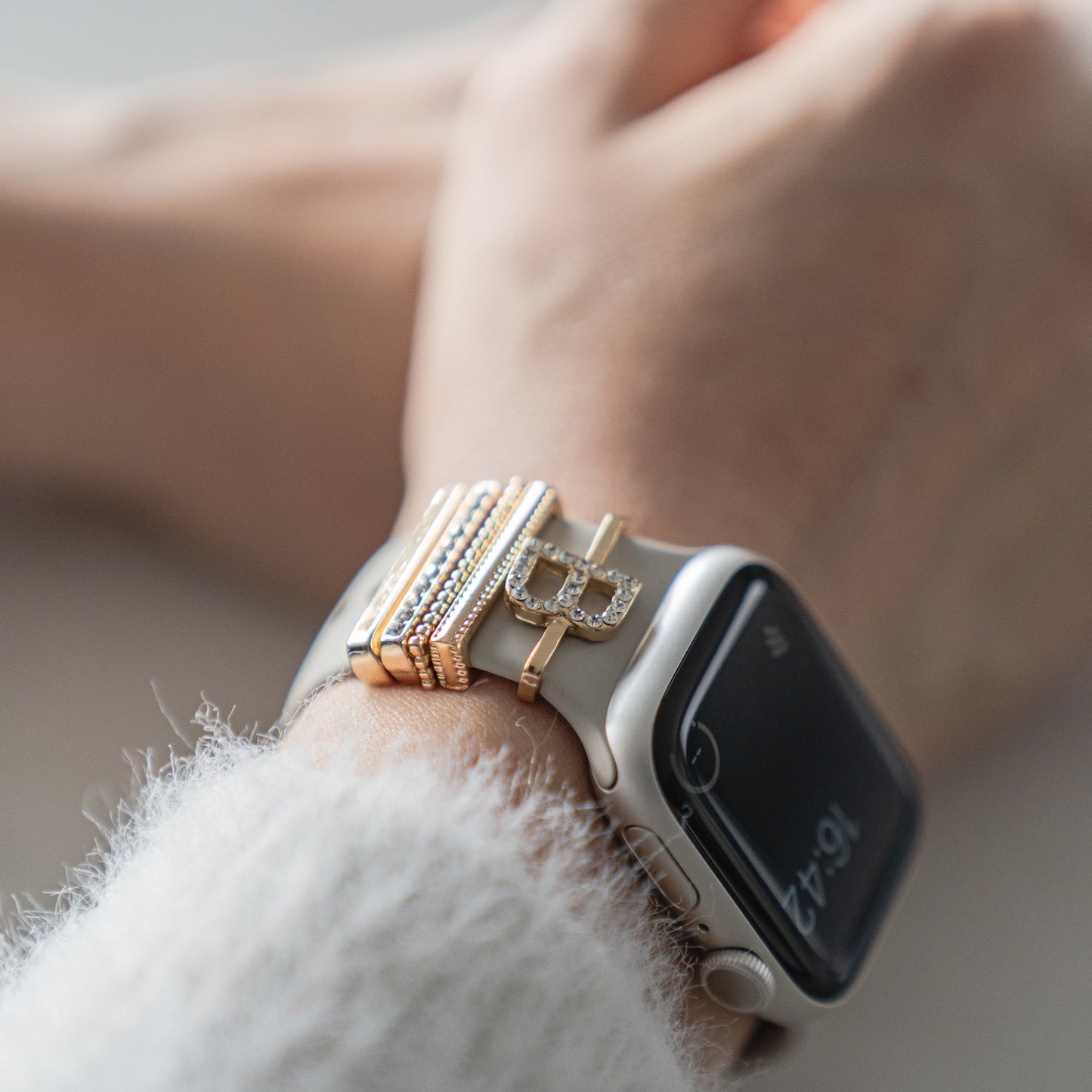 Eine Nahaufnahme eines Handgelenks mit einem arktisband Apple Watch Charms „B Style“ von arktisband auf einem beigen Band, mit goldenen und diamantbesetzten Charms aus Edelstahl und Glas; die Uhr zeigt 10:47.