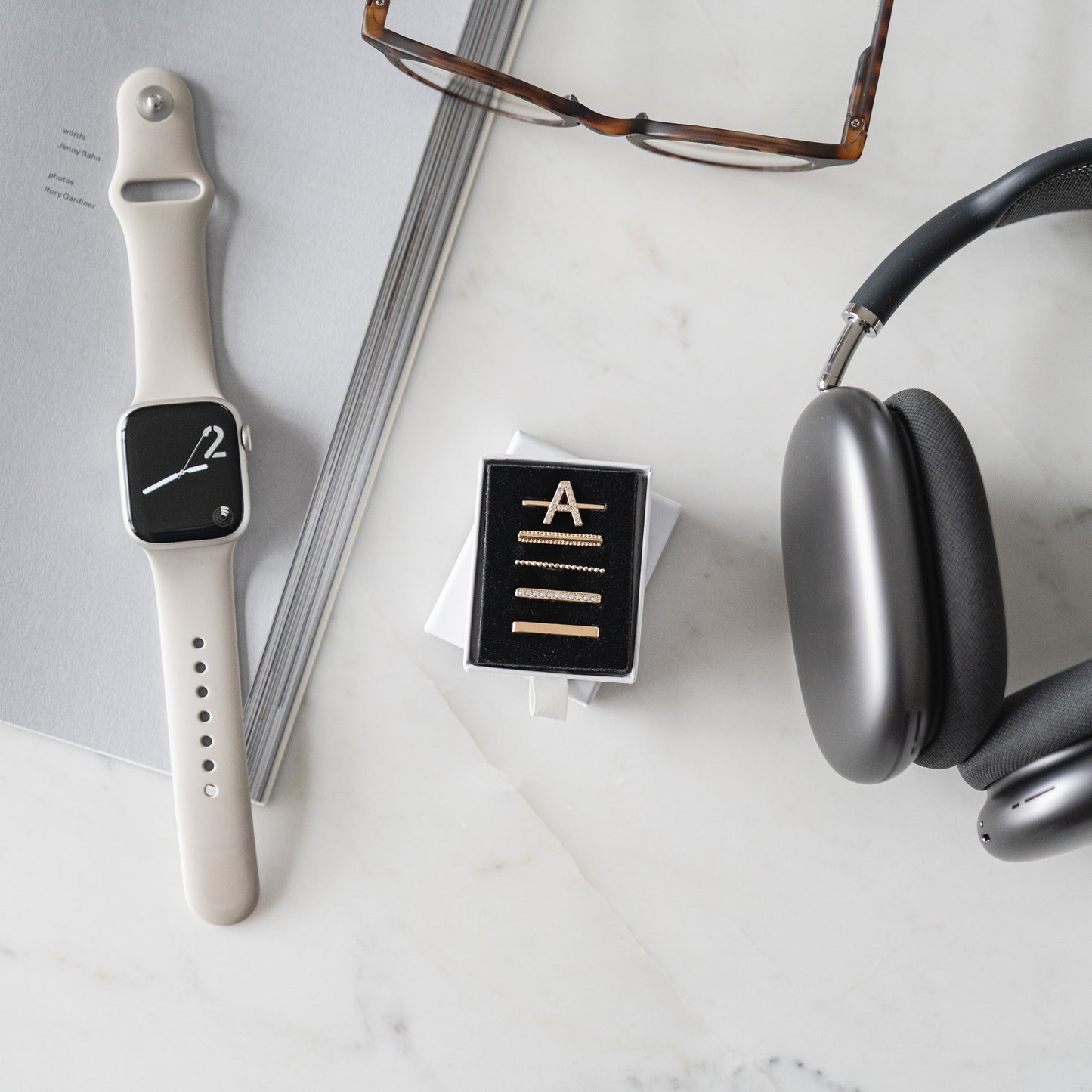 Die arktisband Apple Watch Charms „A Style“ von arktisband glänzen neben Brillen, Haarnadeln in einer Schachtel und Kopfhörern auf einer weißen Marmoroberfläche neben einem grauen Notizbuch – gestylt mit arktisband-Finesse.