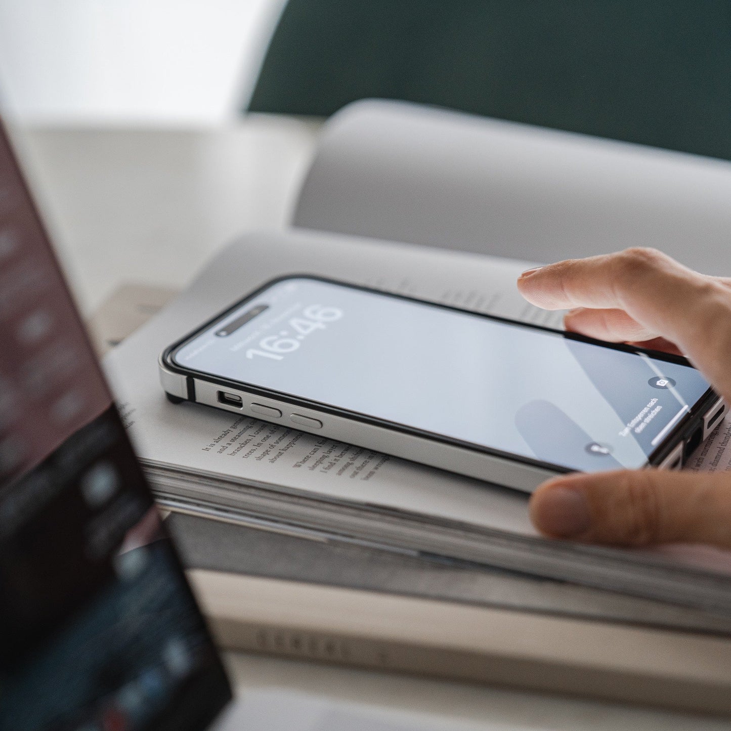 Eine Hand greift nach einem iPhone 15 Plus, geschützt durch den Aviato AirZERO Alu Bumper. Das Display zeigt 16:46, auf einem offenen Buch neben einem Laptop. Diese Aviato-Hülle bietet dank robustem Flugzeugaluminium stoßabsorbierenden Schutz.