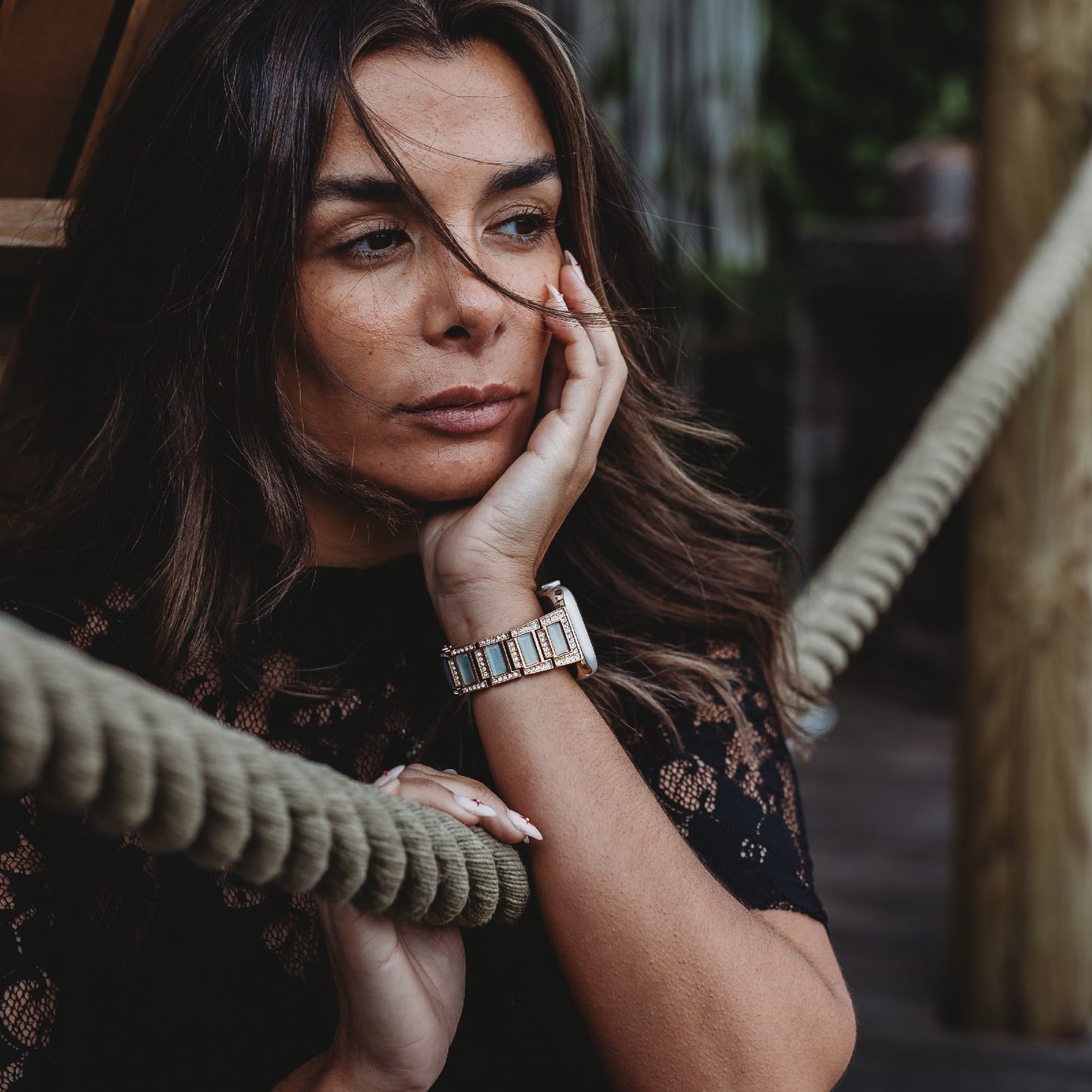 Eine Frau mit langen braunen Haaren, die ein schwarzes Spitzentop und das arktisband Apple Watch Gliederarmband „Senorita“ trägt, ruht im Freien mit dem Gesicht auf ihrer Hand, ihr nachdenklicher Blick wird durch das stylische arktisband Armband unterstrichen.