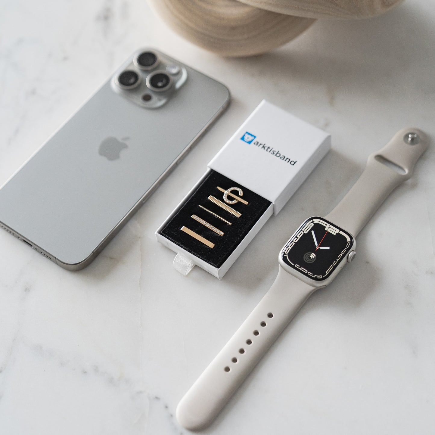 Auf einer weißen Marmoroberfläche sind ein silbernes iPhone, eine beige Apple Watch mit Uhrzeitanzeige und eine Arktisband-Box mit Apple Watch Charms „C Style“ von Arktisband angeordnet.