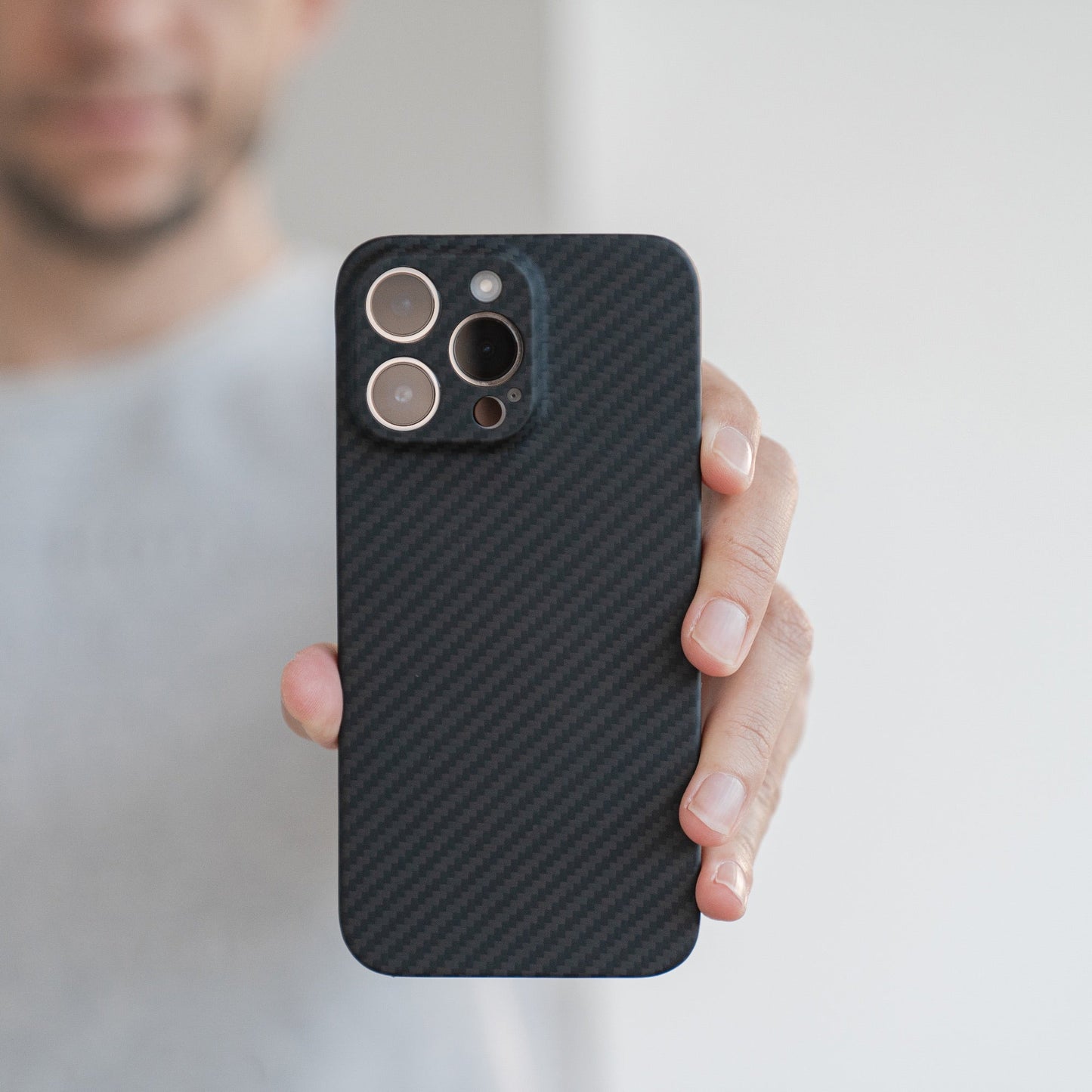 Eine Person hält ein Arktis iPhone 16 Pro Carbon Case aus Carbon- und Aramidfasern in Richtung der Kamera und hebt die MagSafe-Kompatibilität und die markanten Kameraobjektive vor einem schlichten, unscharfen Hintergrund hervor.