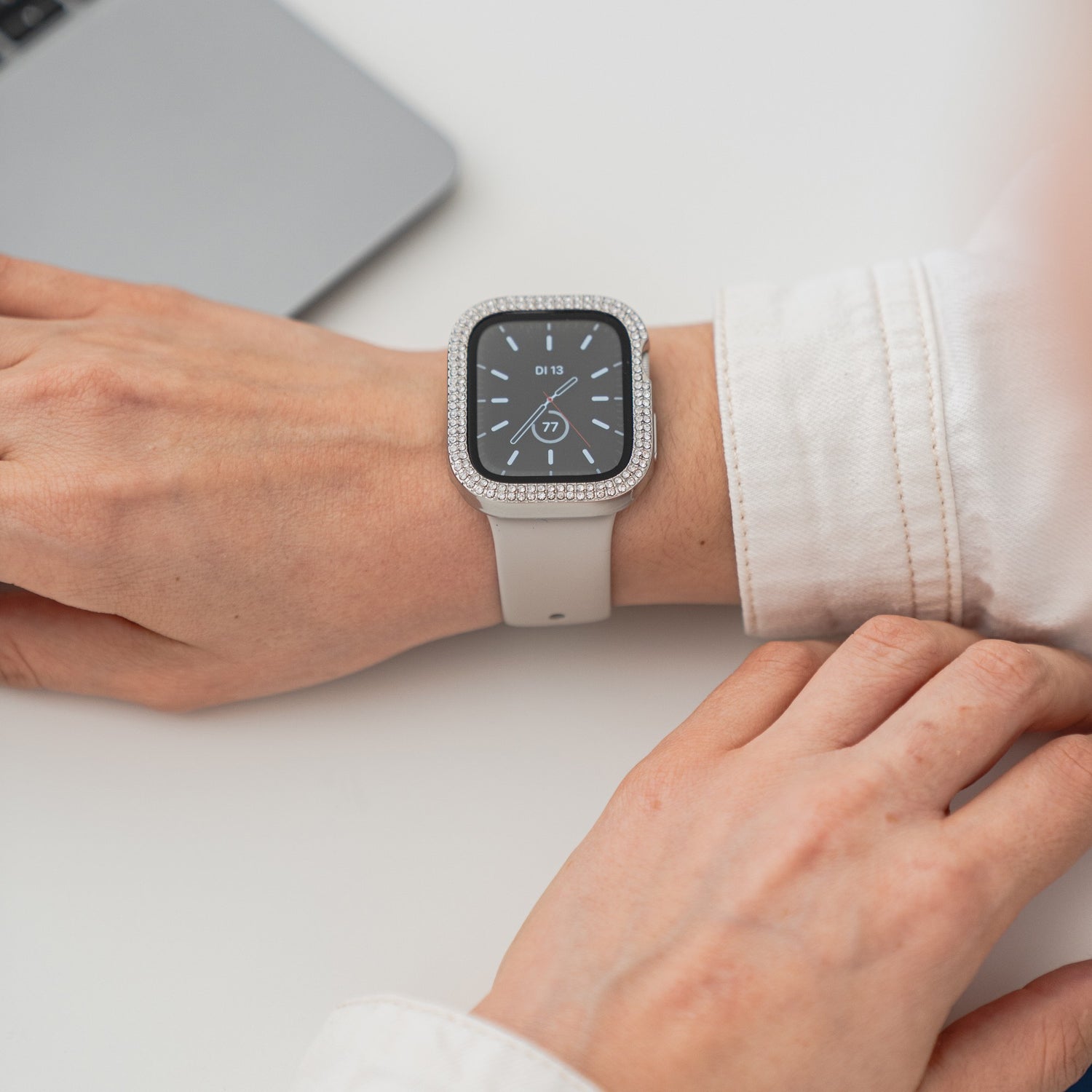 Jemand trägt die arktisband GLAMOUR Glitzerhülle für die Apple Watch mit Strasssteinchen auf der Lünette und weißem Armband. Die Hand ruht auf einer weißen Fläche neben einem silbernen Laptop, auf dessen Zifferblatt 17:19 Uhr angezeigt wird.