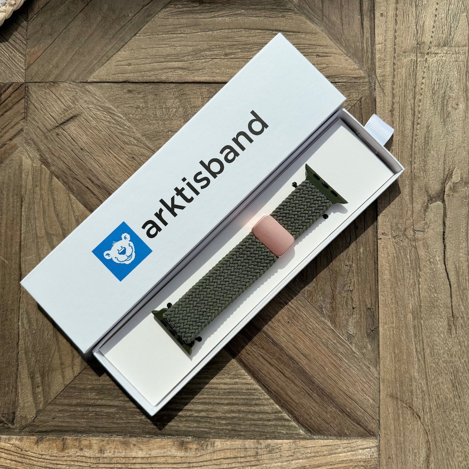 Ein grünes Armband Geflochtenes Apple Watch Armband „Casual“ mit Magnetverschluss und pinker Schlaufe, präsentiert in der Armband-Box auf einer Holzoberfläche.