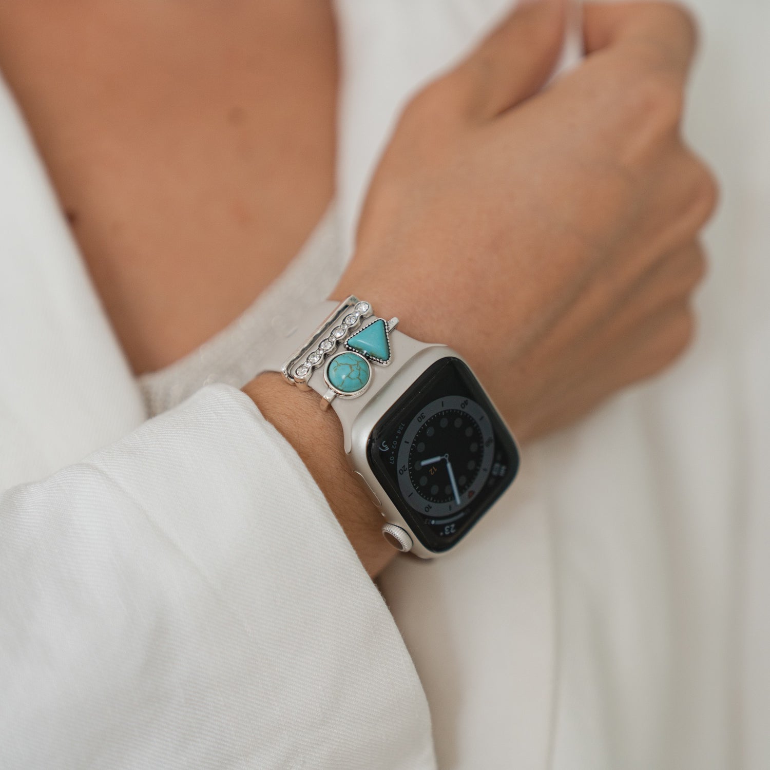 Eine Person in einem weißen Ärmel trägt Arktisband Apple Watch Charms „Opal“ – Silber und ein silbernes Armband mit türkisfarbenen Steinen, die Edelstahl und Glas für einen stilvollen Look kombinieren.