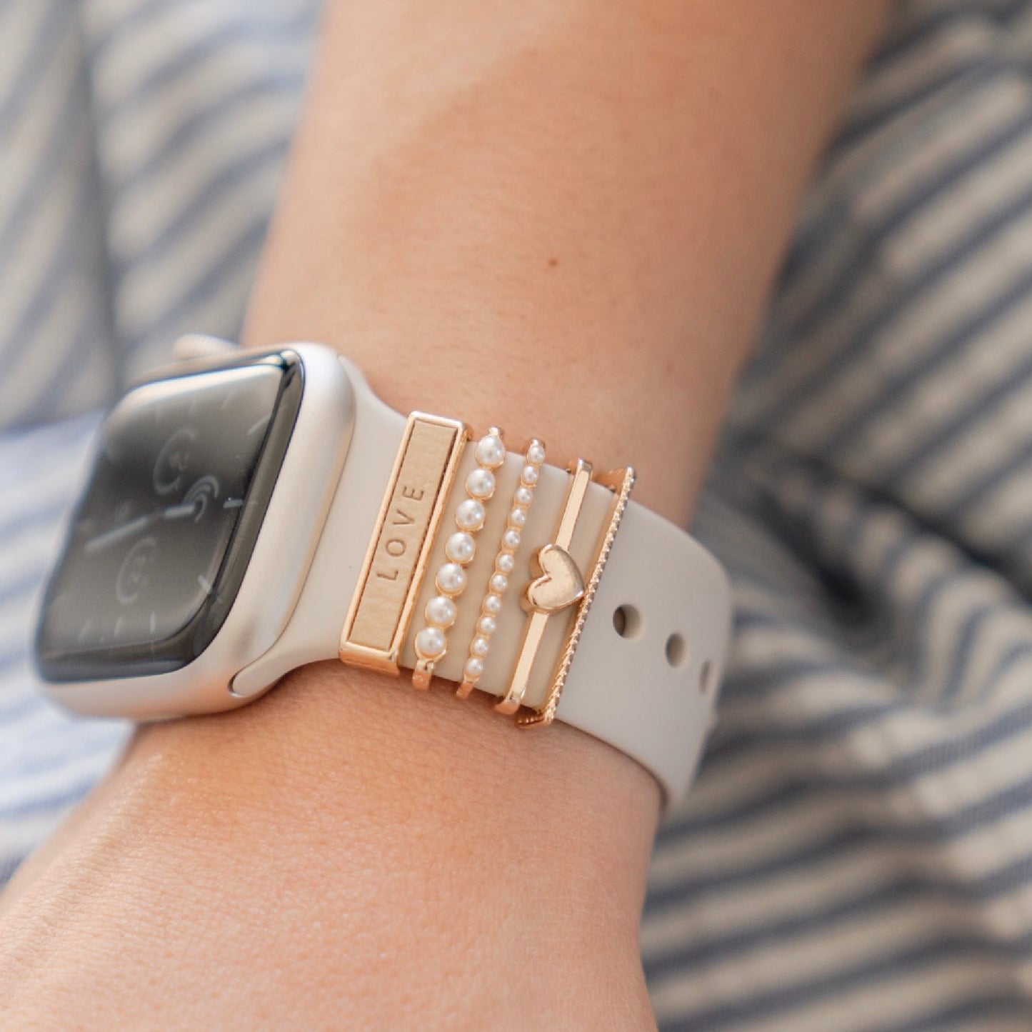 Eine weiße Arktisband-Smartwatch mit Apple Watch Charms „Love“ in Gold und Perle, versehen mit einem „LOVE“- und Herz-Charm von Arktisband, wird an einem Handgelenk vor blau-weiß gestreiftem Stoff präsentiert.