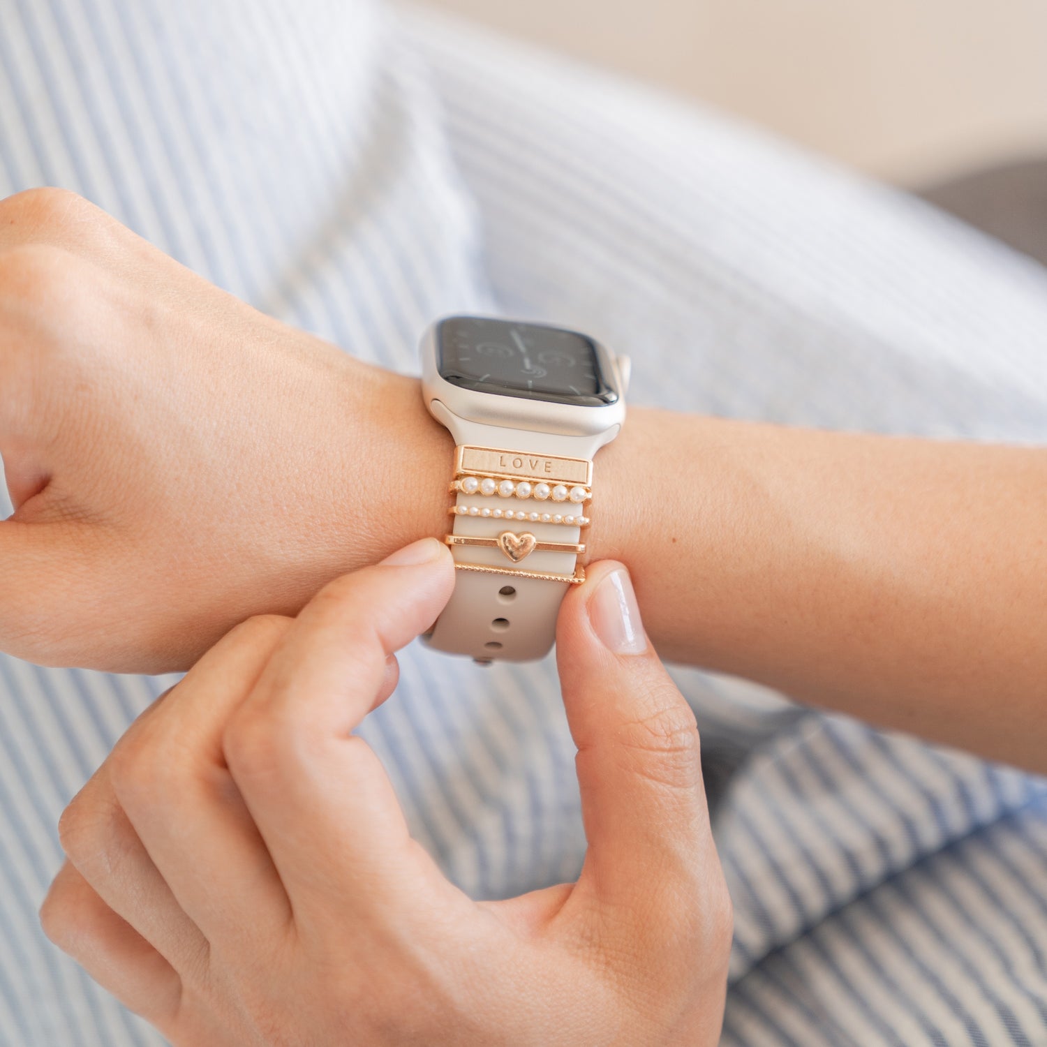Eine Person passt ein beiges Arktisband-Smartwatch-Armband mit goldenen Arktisband-Apple-Watch-Charms „Love“ und einem Herz an, im Hintergrund ist ein blau-weiß gestreifter Stoff zu sehen.