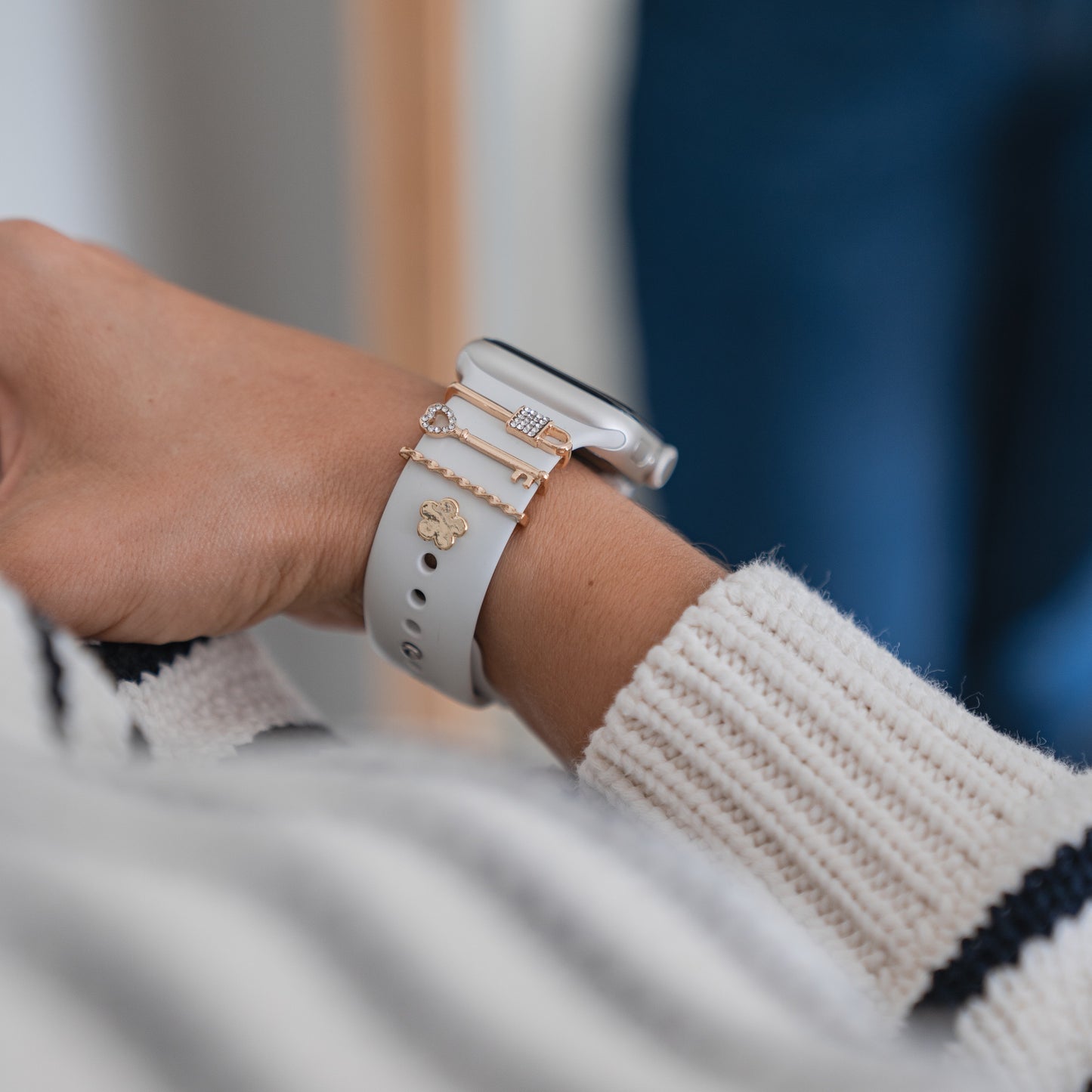Nahaufnahme eines Handgelenks mit Arktisband-Apple-Watch-Charms „Golden Key“ von Arktisband, kombiniert mit anderen Charm-Accessoires, einem weißen Strickpullover und einem Arktisband aus Edelstahl und Glas.