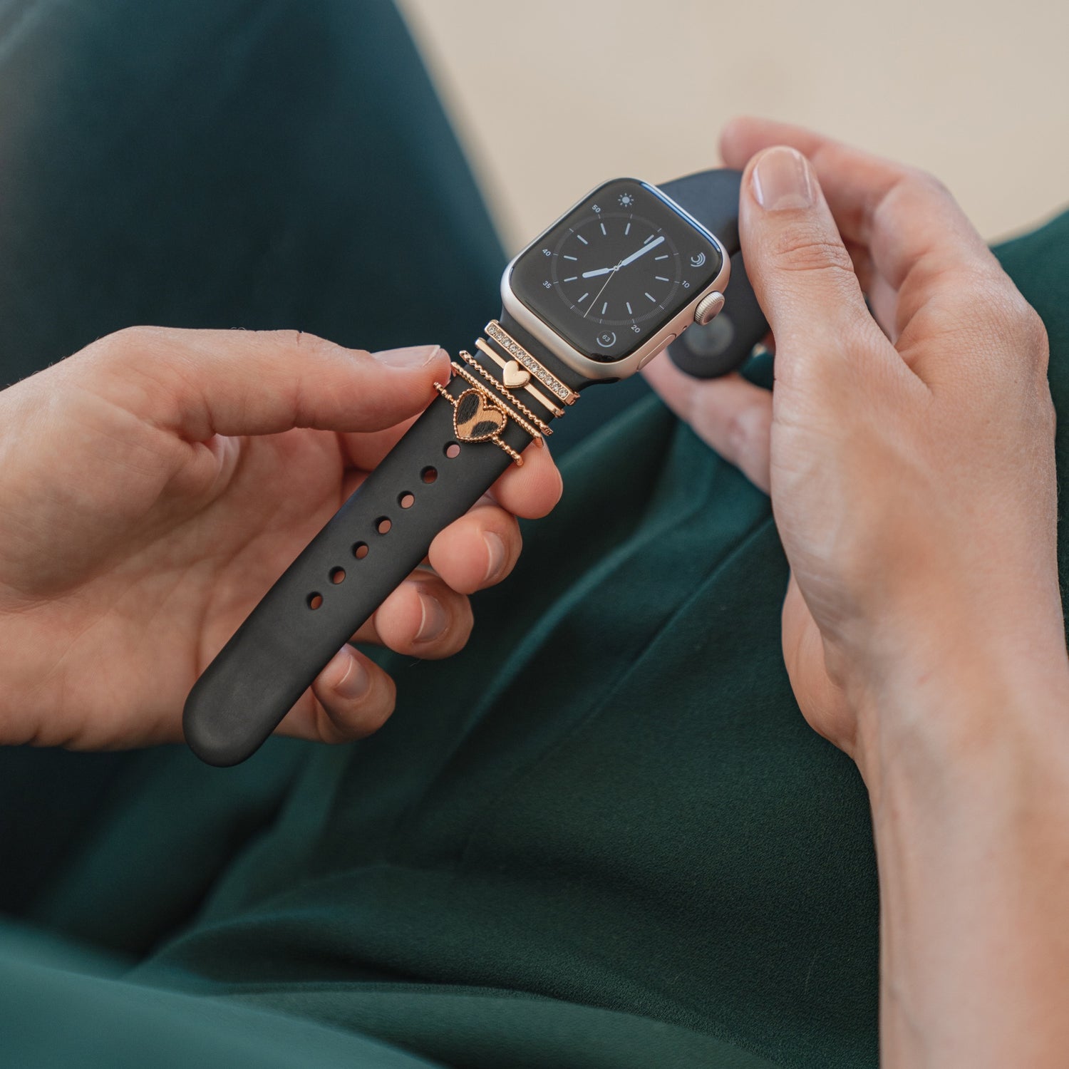 Eine Person in grüner Kleidung hält eine Apple Watch mit einem schwarzen Arktisband und goldenen Arktisband-„Herz“-Anhängern auf ihrem Schoß.