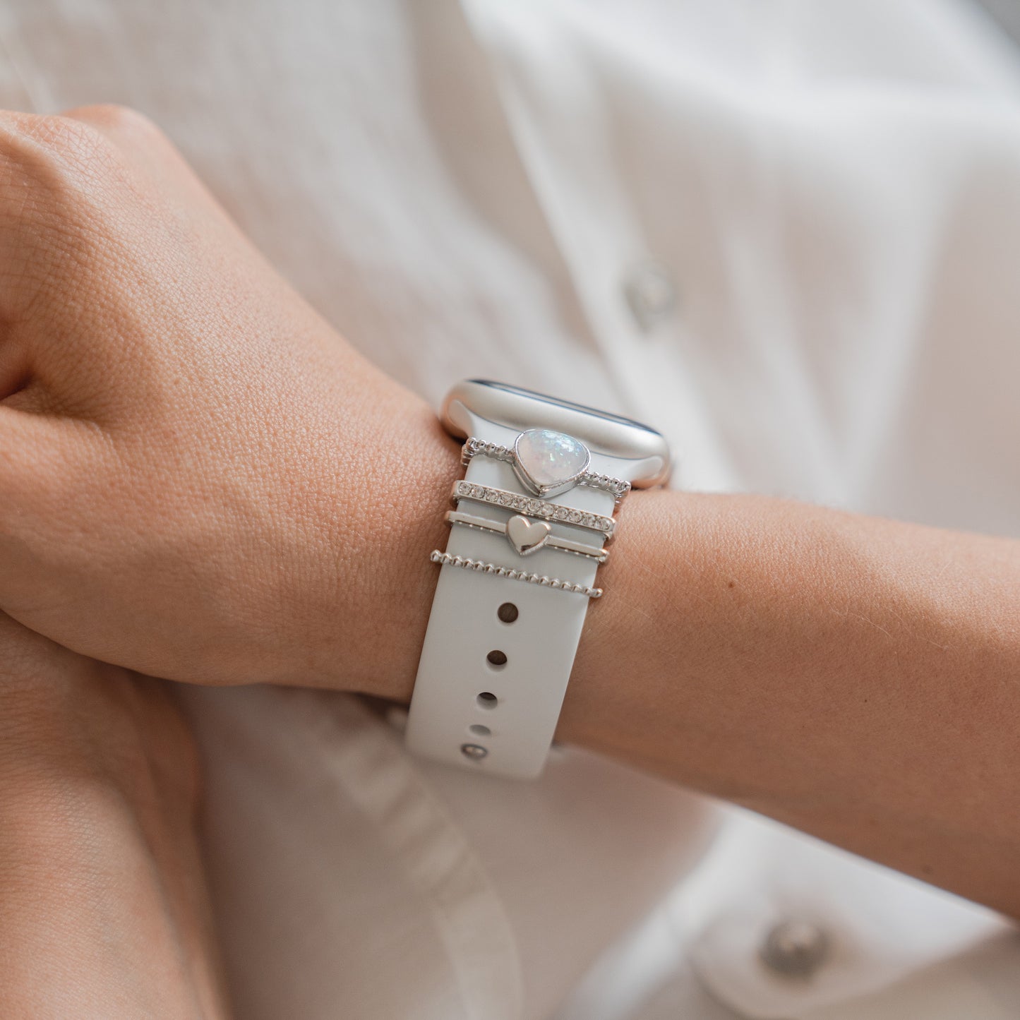 Am Handgelenk einer Person schmückt das Arktisband Apple Watch Charms „Loyal“ – Silber, geschmückt mit silbernen und juwelenbesetzten Ringen sowie einem Herzanhänger, perfekt kombiniert mit einem frischen weißen Hemd für einen stylischen Look.