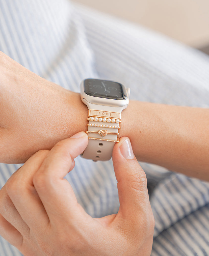 Eine Hand trägt eine Smartwatch mit dekorativen goldenen Anhängern mit der Aufschrift „LOVE“ und einem Herz auf dem Armband.