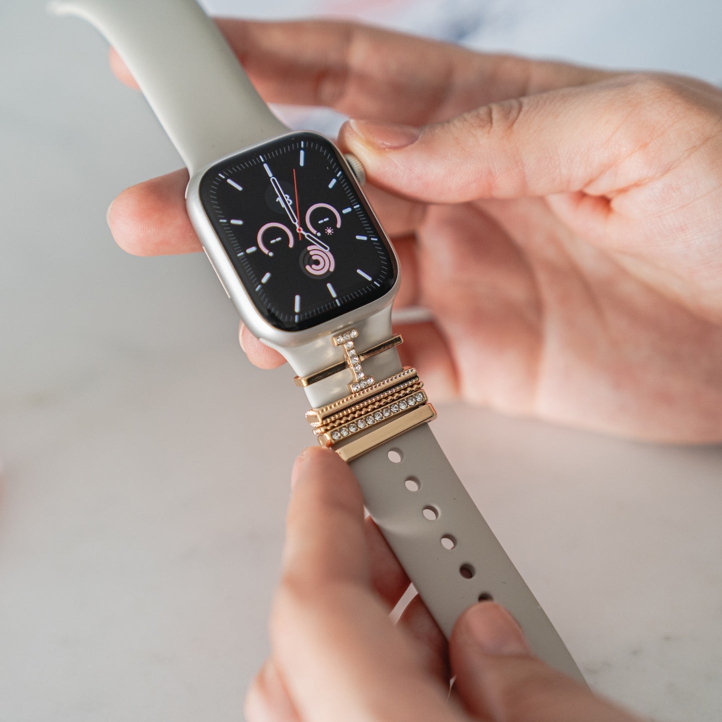 Die Hände einer Person legen Arktisband Apple Watch Charms „I Style“ auf das Armband einer grauen Smartwatch mit Digitalanzeige.