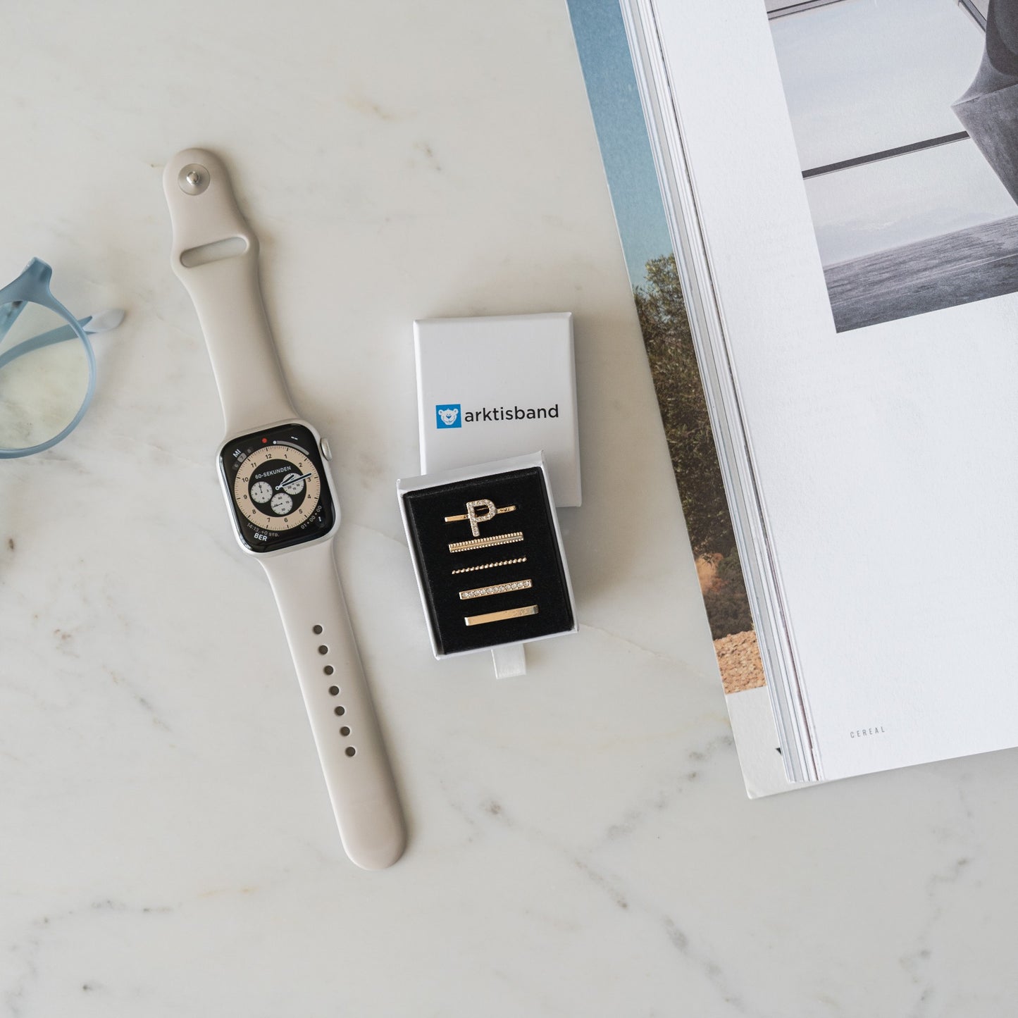Eine beige Smartwatch, Arktisband Apple Watch Charms „P Style“, eine blaue Brille und eine offene Zeitschrift sind auf einer weißen Marmoroberfläche angeordnet.