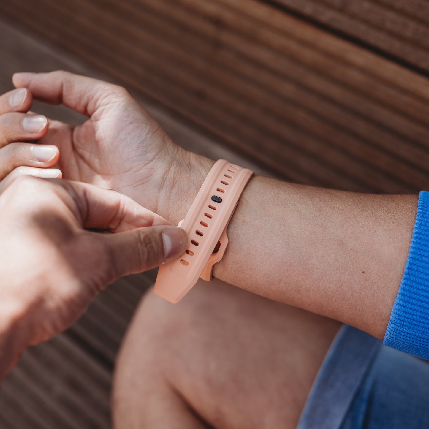 Eine Person befestigt ein Arktisband Apple Watch FKM Sportarmband „Sweat“ in Hellrosa an ihrem Handgelenk. Sie trägt ein blaues Oberteil und Jeansshorts und sitzt auf einer Holzfläche.