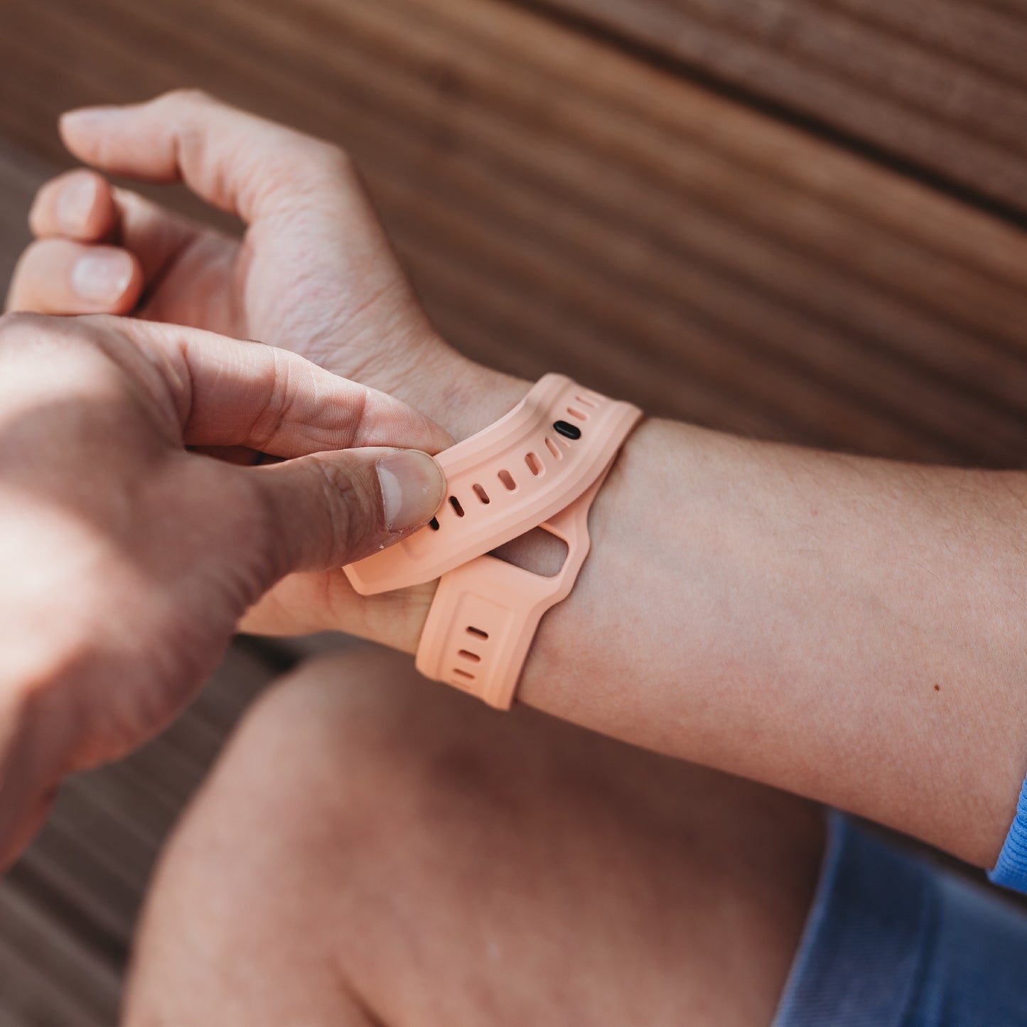 Eine Person befestigt das arktisband Apple Watch FKM Sportarmband "Sweat" in Pfirsichfarbe um ihr Handgelenk, während sie auf einer Holzoberfläche sitzt. Das Armband von arktisband ist robust und flexibel und eignet sich perfekt für den Alltag.