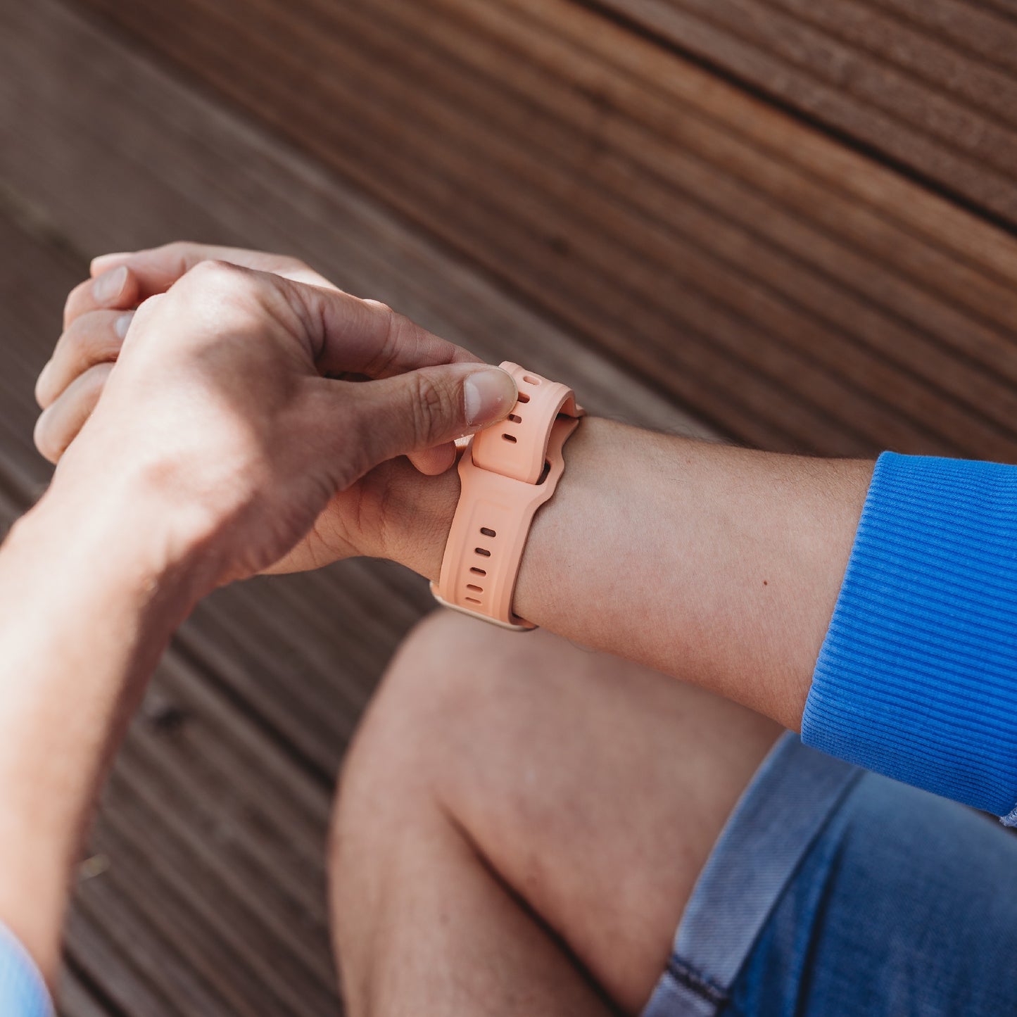 Eine Person in blauem Hemd und Jeansshorts befestigt das arktisband Apple Watch FKM Sportarmband „Sweat“ von arktisband, ein pfirsichfarbenes, strapazierfähiges und elastisches Fluorelastomer-Armband, an ihrem linken Handgelenk. Im Hintergrund sind Holzlatten zu sehen.