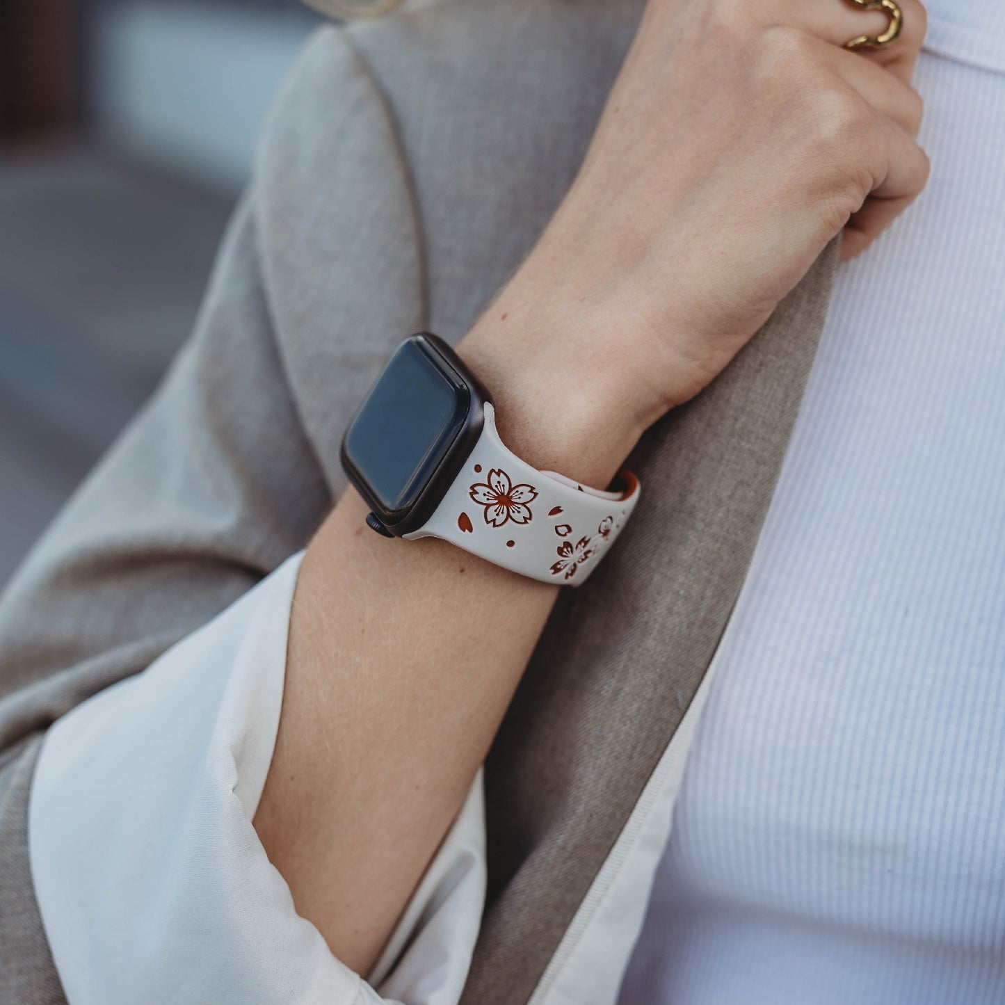 Eine Person in einer beigen Jacke und einem weißen gerippten Oberteil trägt das arktisband Apple Watch Armband „Flores“ von arktisband mit einem schweißabsorbierenden Blumenmuster, gepaart mit einem goldenen Ring am Finger.