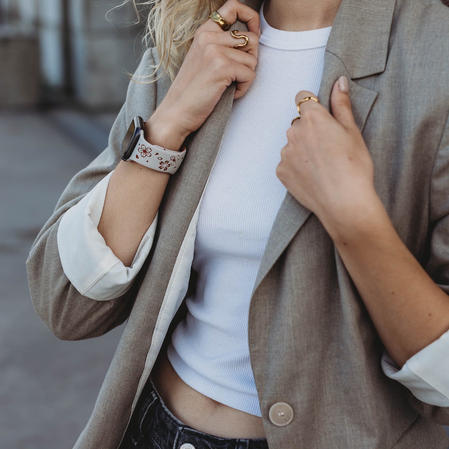 Eine Person mit welligem blondem Haar, mehreren Ringen und einem Arktisband-Apple-Watch-Armband „Flores“ rückt das Revers eines hellbraunen Blazers zurecht, der über einem weißen gerippten bauchfreien Top und dunklen Jeans getragen wird.