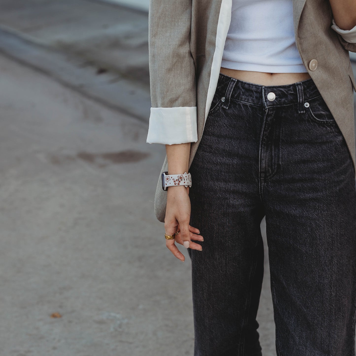 Eine Person in einem beigen Blazer mit hochgekrempelten Ärmeln, einem weißen Tanktop und schwarzen Jeans mit hoher Taille steht auf Beton und zeigt Ringe und das arktisband Apple Watch Armband „Flores“ von arktisband an ihrem linken Handgelenk.