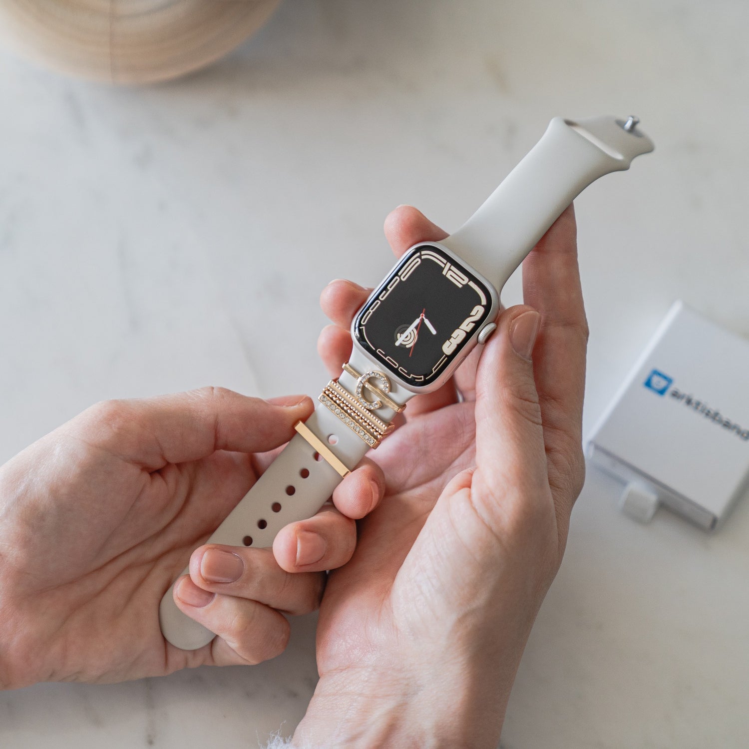 Eine Person passt das Armband einer Apple Watch mit weißem Band und goldenem Anschluss an und fügt möglicherweise einen Arktisband Apple Watch Charm „C Style“ hinzu. Im Hintergrund steht eine kleine weiße Box mit blauer Schrift auf dem Tisch.