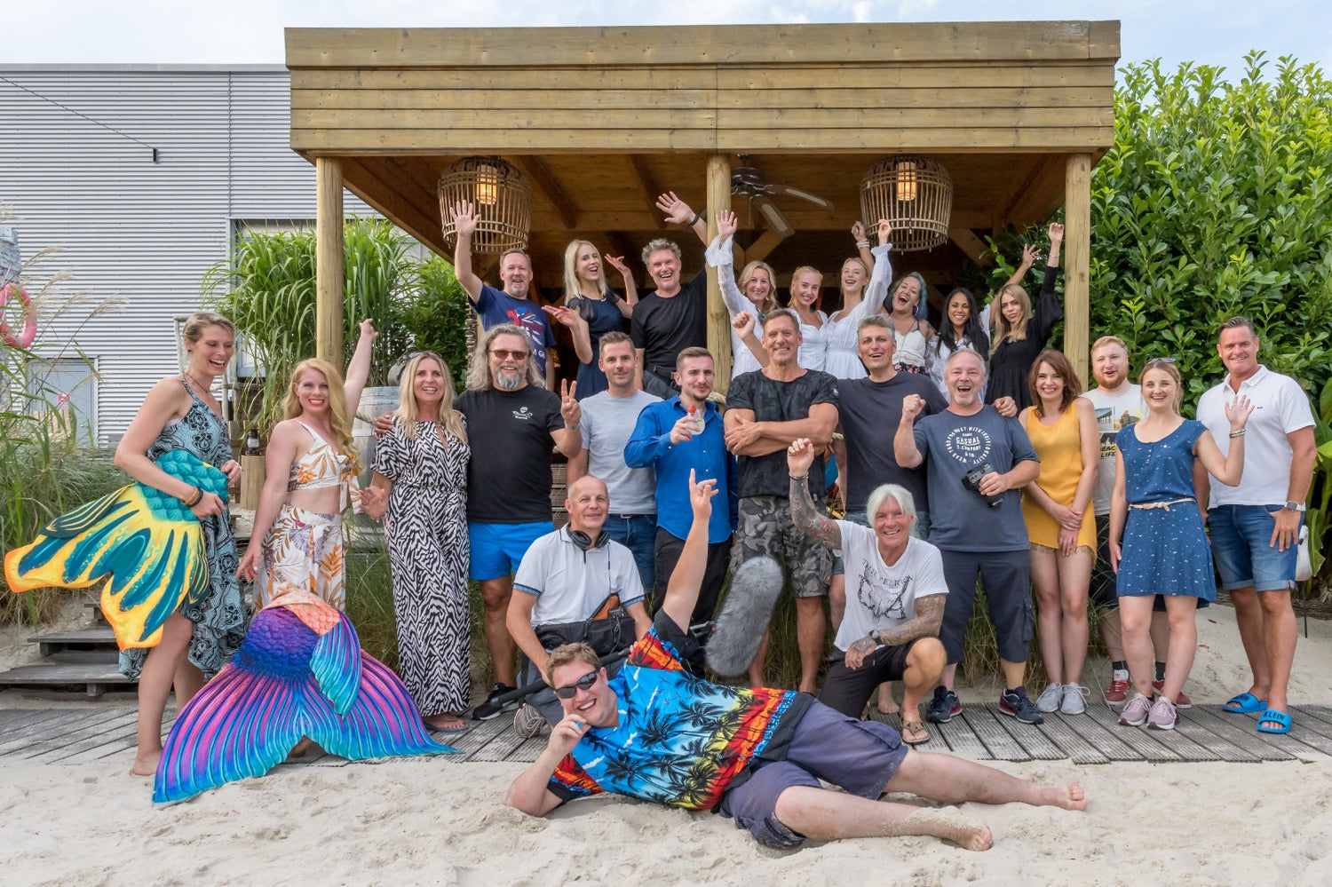 Eine Gruppe Erwachsener posiert und lächelt gemeinsam an einem Sandstrand vor einer Holzhütte.