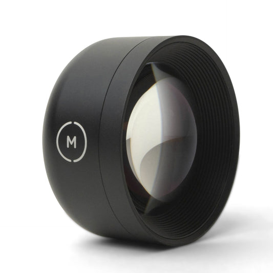 Das MOMENT 58mm Tele Lens T-Series, ein schwarzer Kameraobjektivaufsatz mit M-Logo, platziert auf einer weißen Oberfläche, bietet optische Vergrößerung bei der Verwendung als iPhone Teleobjektiv.