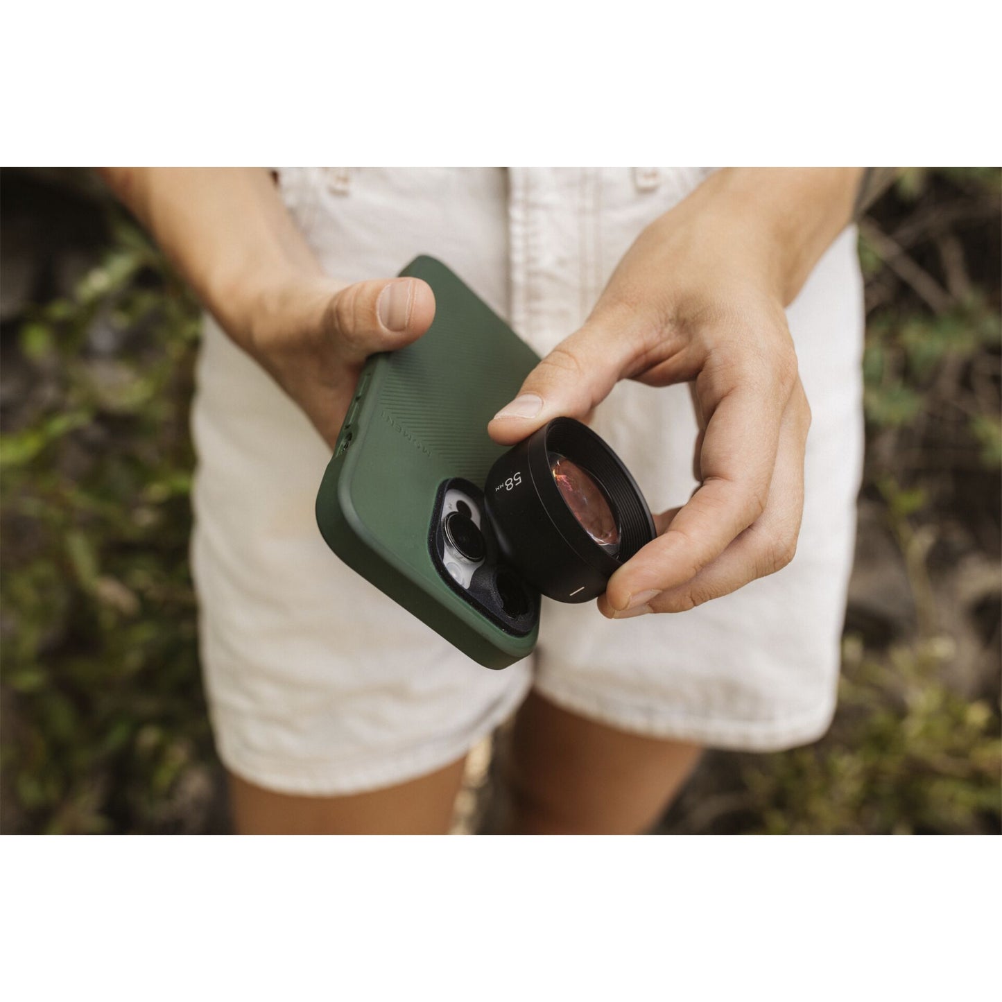 Hände, die das Moment Tele Lens 58mm T-Series von MOMENT an einer grünen Handyhülle befestigen, wobei die Person weiße Shorts trägt.