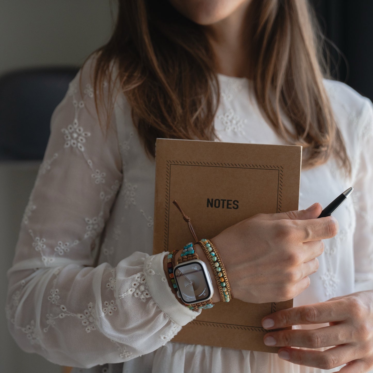 Eine Frau in einem weißen Spitzenkleid hält ein Notizbuch und einen Stift in der Hand und trägt dazu bunte Armbänder, eine Uhr und das arktisband Apple Watch Armband "Dream Catcher" von arktisband für einen schicken Touch.