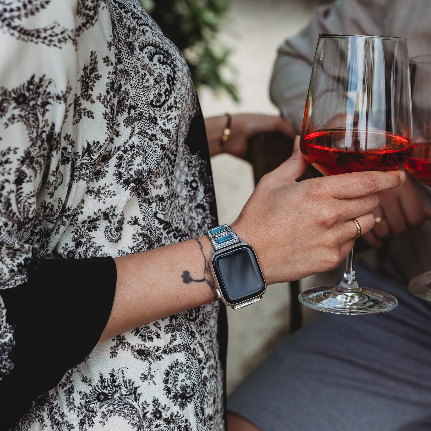 Eine Person, die ein gemustertes weiß-schwarzes Oberteil, ein Arktisband Apple Watch Gliederarmband „Senorita“ und ein Perlenarmband trägt, hält ein Glas Rotwein, während eine andere Person im Hintergrund ebenfalls Rotwein genießt.
