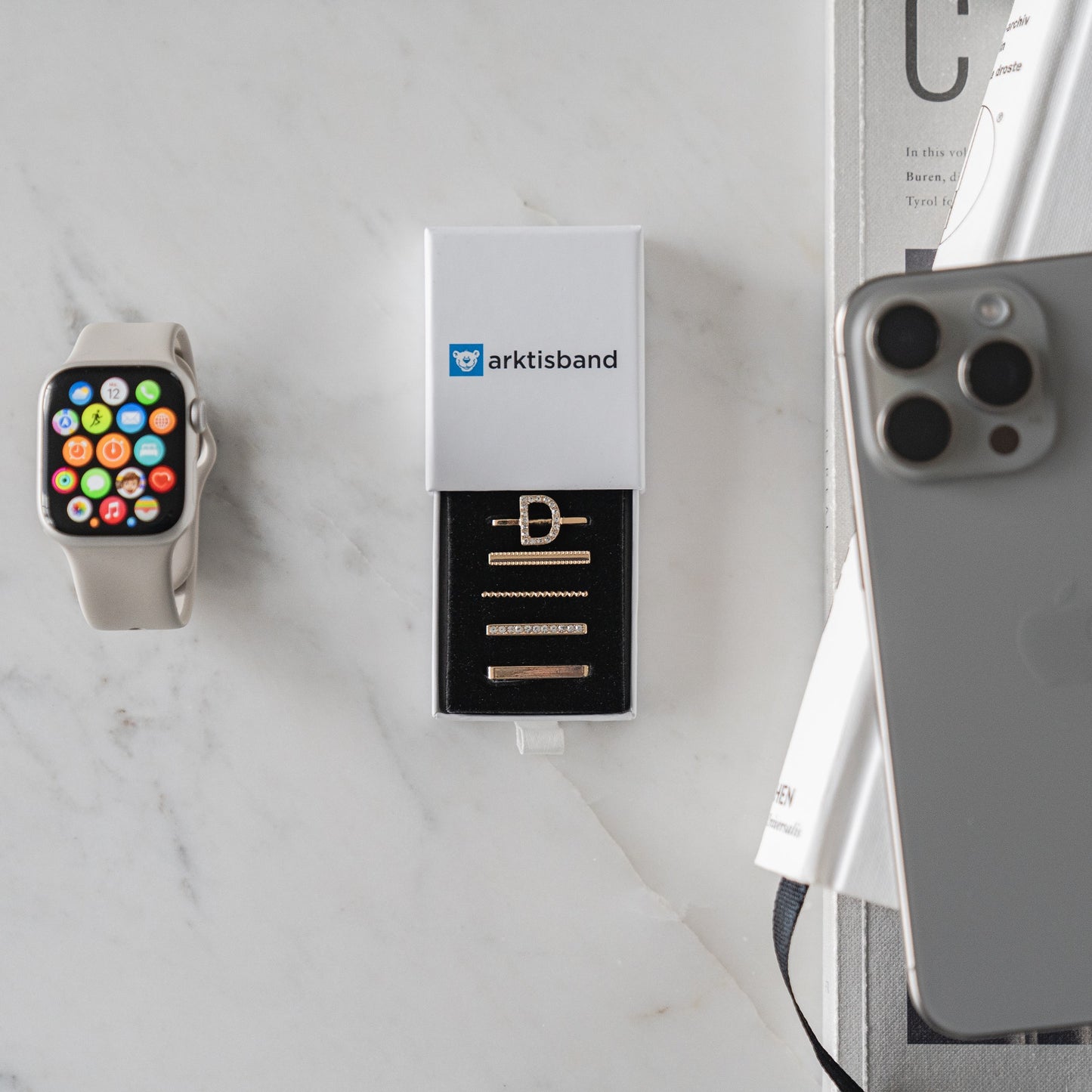 Apple Watch mit Arktisband Apple Watch Charms „D Style“ und einem iPhone auf einer Marmoroberfläche, Zeitschrift in der Ecke.