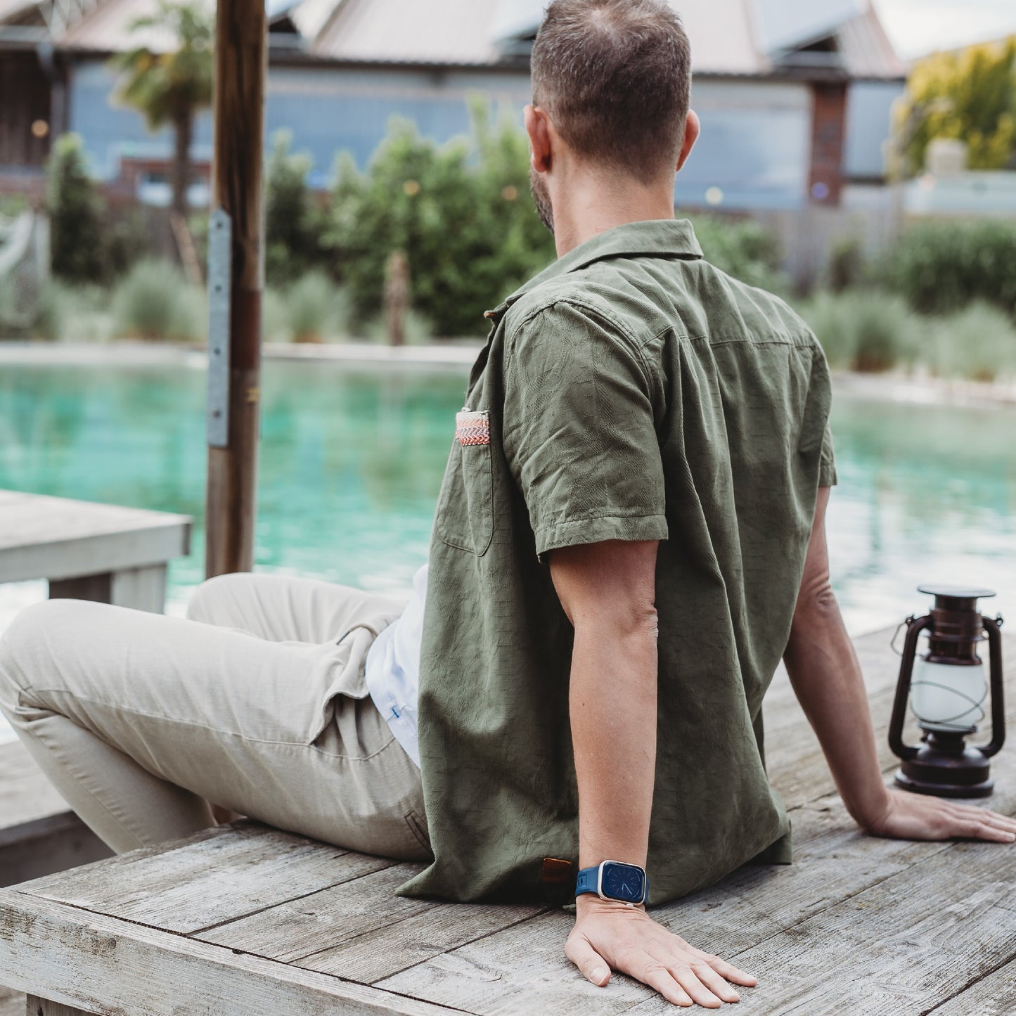Ein Mann in olivgrünem Hemd und beiger Hose sitzt mit abgewandtem Blick auf einem Holztisch neben einem Pool. Er trägt das arktisband Apple Watch FKM Sportarmband „Sweat“ von arktisband. In der Nähe des Tisches steht eine altmodische Laterne.