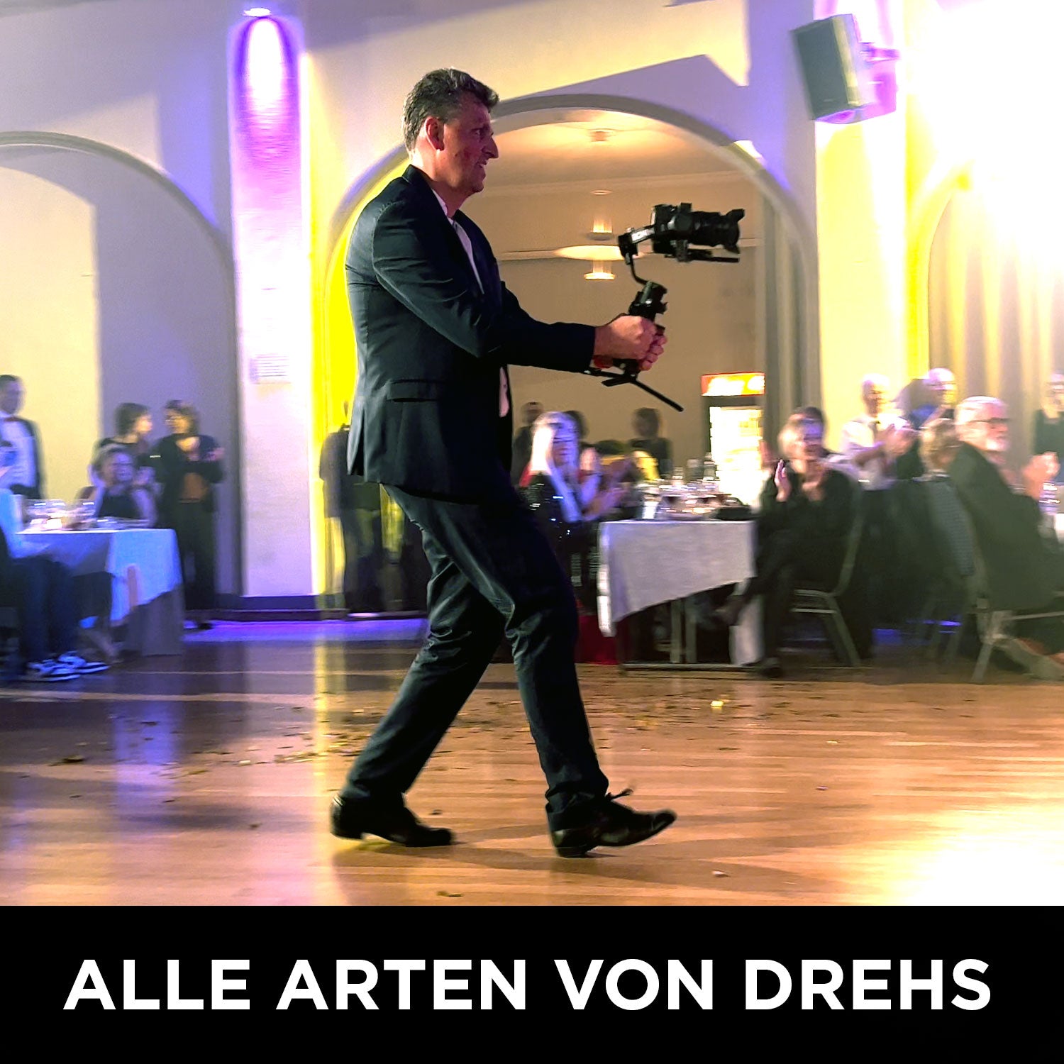 Ein Mann im Anzug geht über einen Holzboden und filmt mit einem Stabilisator, während das Publikum unter violettem Licht sitzt. Der Text lautet: ALLE ARTEN VON DREHS – Einfach filmen lernen mit dem Wolffilms Academy Online-Kurs von Rainer Wolf.