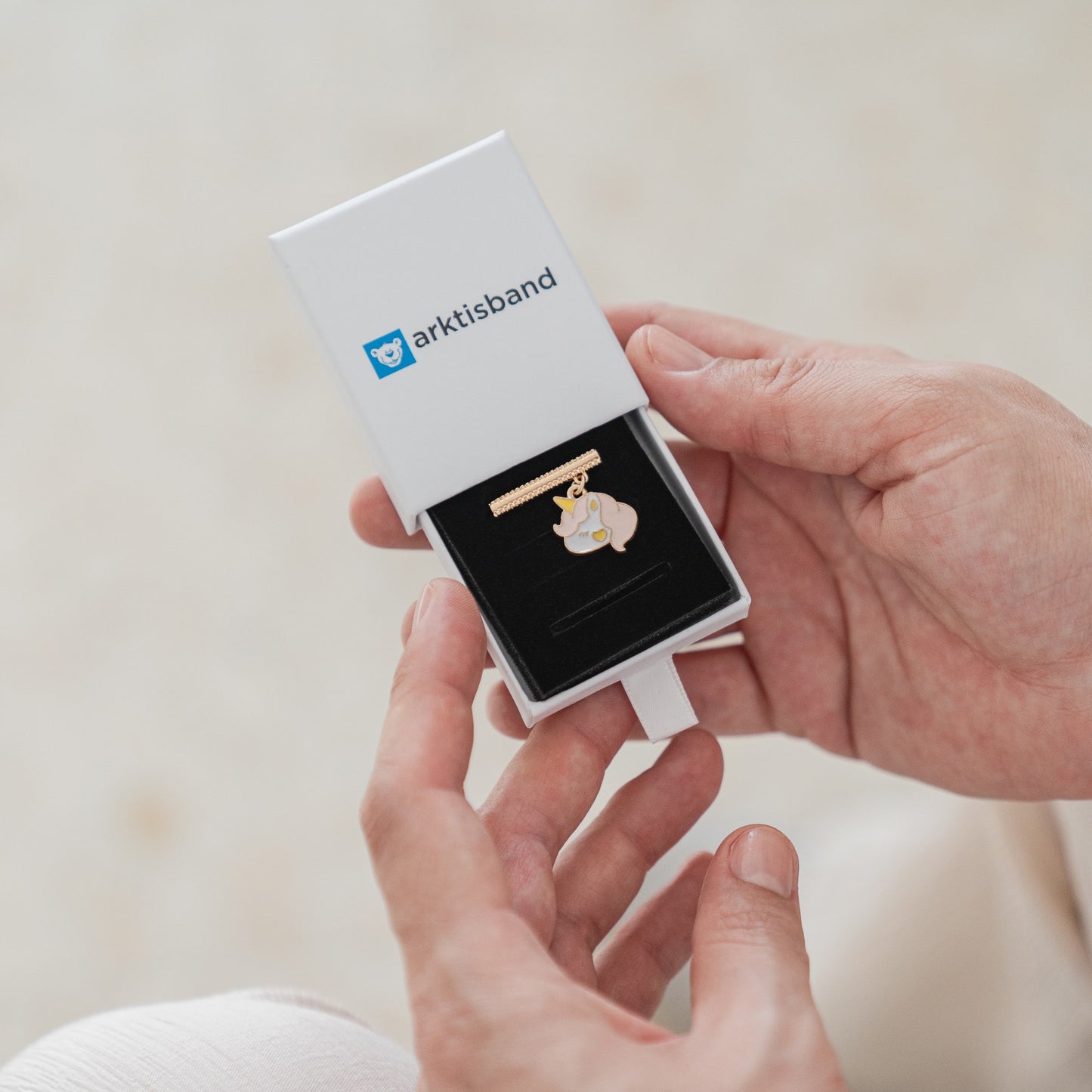 Eine Person hält eine geöffnete weiße Box mit dem Arktisband-Logo in der Hand. Darin befindet sich ein Arktisband-Armband für die Apple Watch in Form eines Eisbären. Ihre Hände und ein Teil ihrer cremefarbenen Hose sind ebenfalls zu sehen.