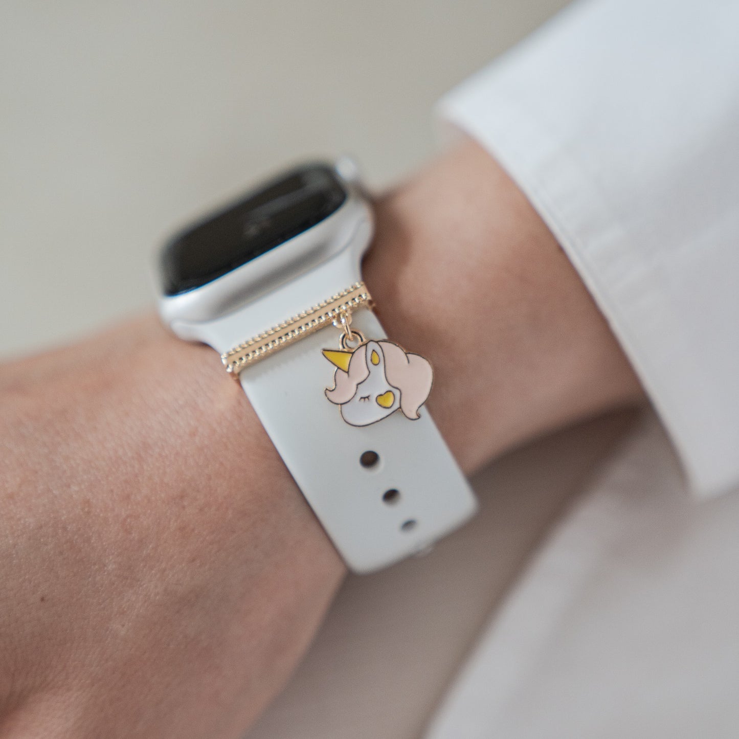 Eine Person in einem langärmeligen weißen Hemd trägt eine weiße Smartwatch, die mit einem gold-rosa Einhorn-Anhänger aus der Arktisband Apple Watch Armband Charms-Kollektion von Arktisband verziert ist.