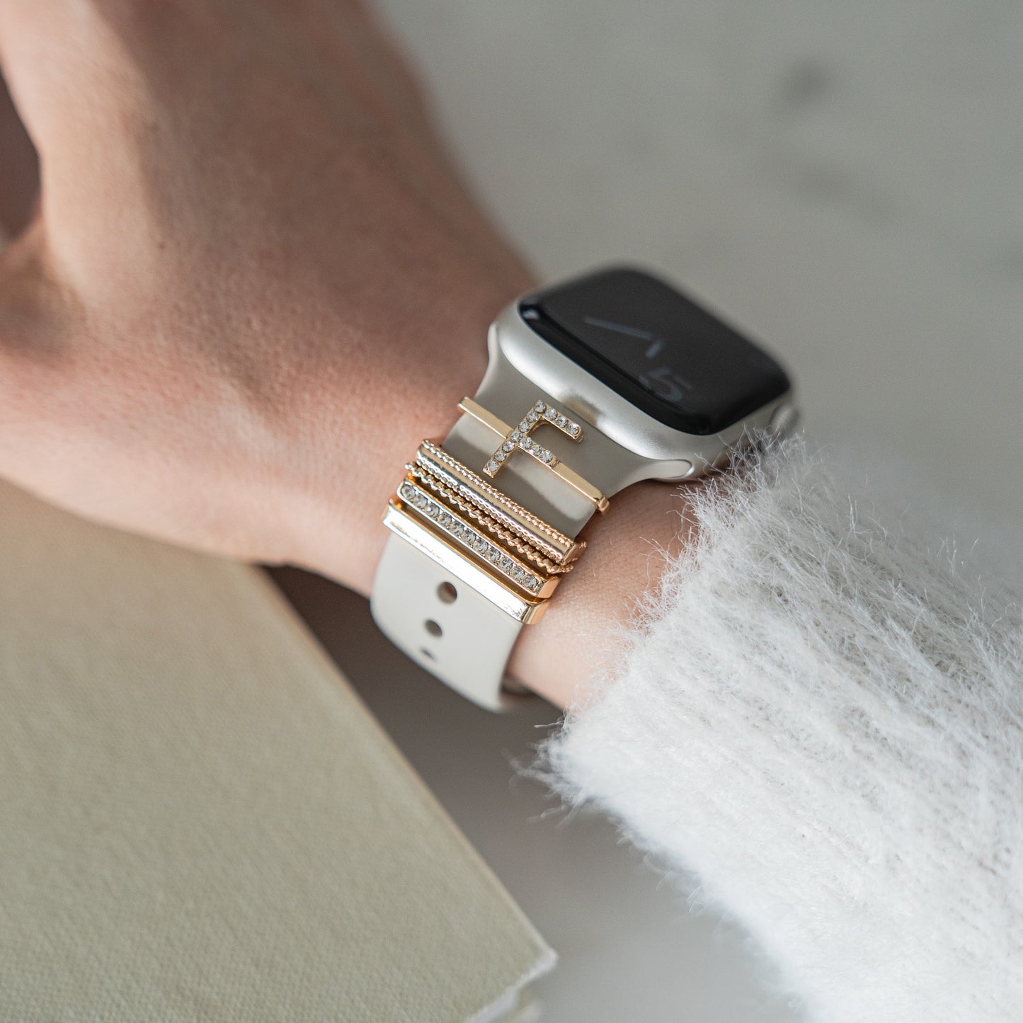 Ein Handgelenk mit arktisband Apple Watch Charms „F Style“ und gestapelten goldenen und diamantenen Buchstaben- und Band-Charms von arktisband auf einem weißen Armband.