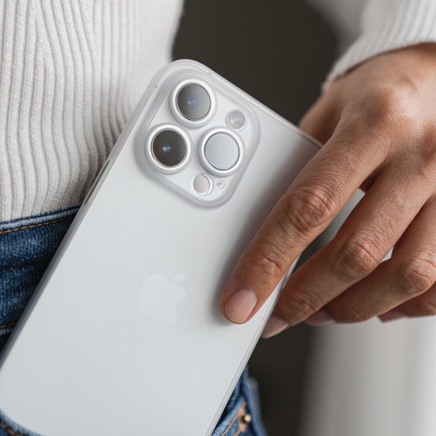 Eine Person schiebt ein weißes iPhone 15 mit drei Kameraobjektiven, geschützt durch die ArktisPRO iPhone 15 Hülle ULTRADÜNN, in die Gesäßtasche einer Bluejeans, während sie einen weißen Rippenpullover trägt.