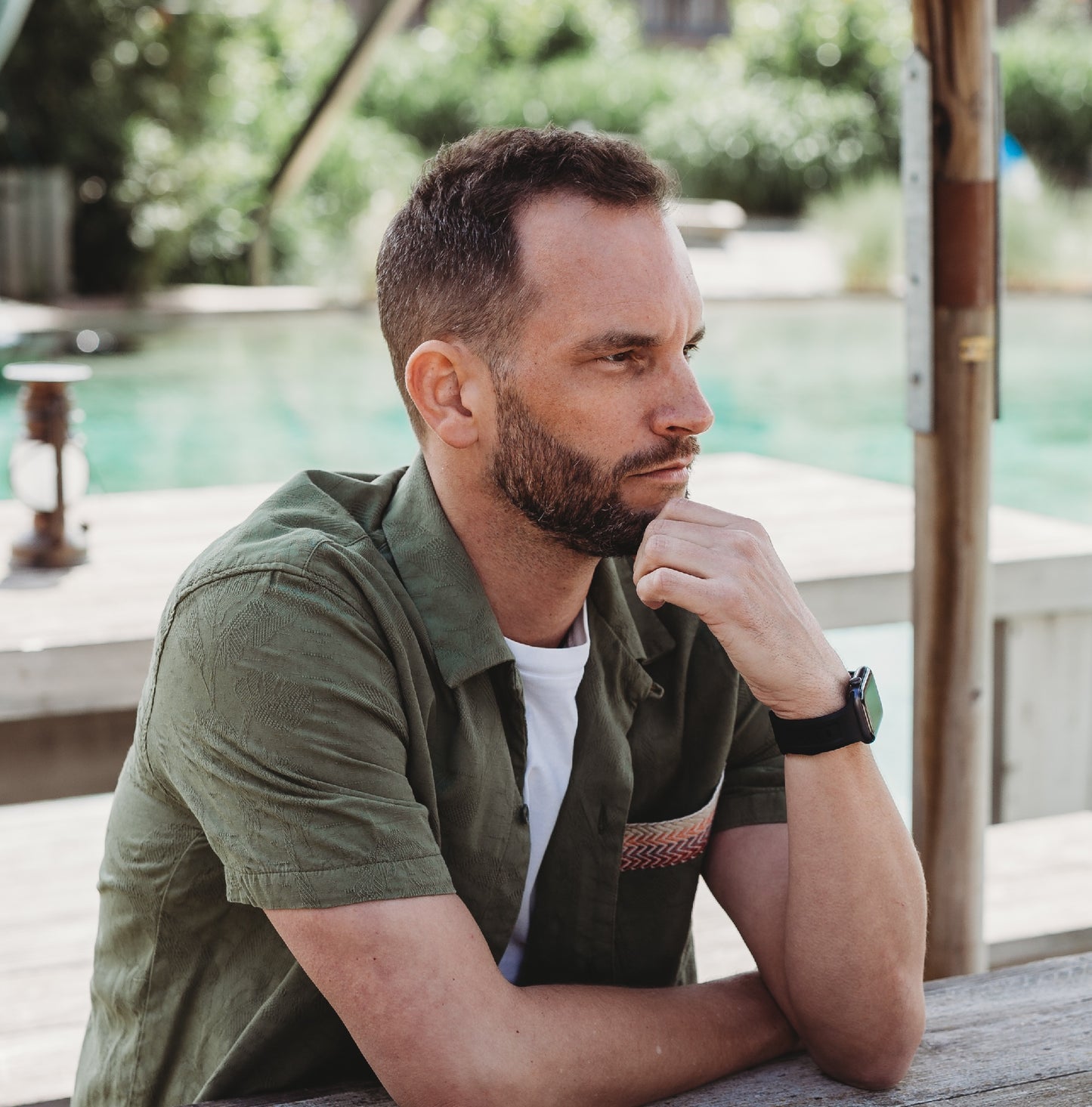 Ein Mann mit kurzen braunen Haaren und Bart sitzt im Freien an einem Holztisch und stützt sein Kinn auf seine Hand. Er trägt ein grünes Hemd, ein weißes T-Shirt und das arktisband Apple Watch FKM Sportarmband „Sweat“ von arktisband.