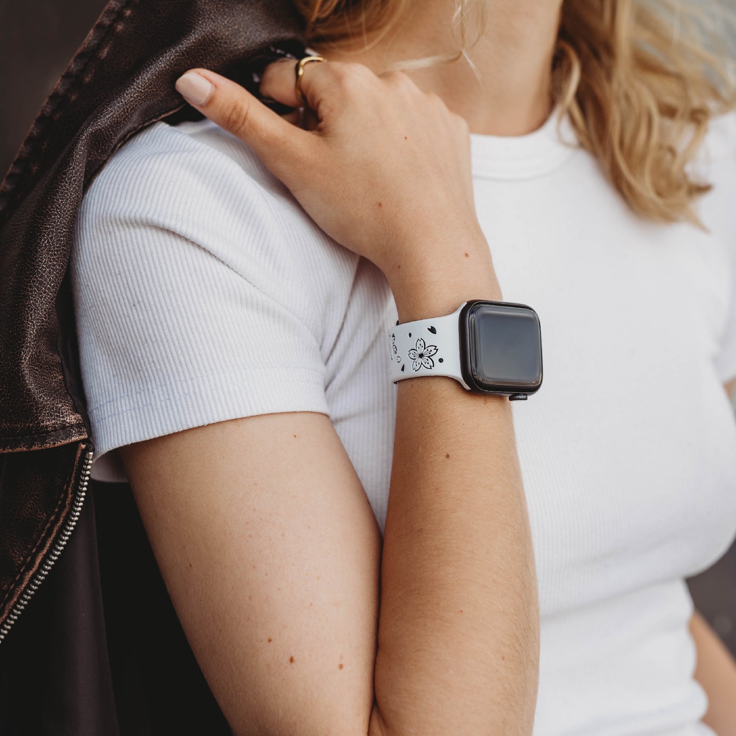 Eine Person trägt ein weißes geripptes Kurzarmhemd und ein weißes Arktisband-Apple-Watch-Armband „Flores“ von Arktisband, mit ihrer Apple Watch (leerer Bildschirm) und einer braunen Jacke über der Schulter.