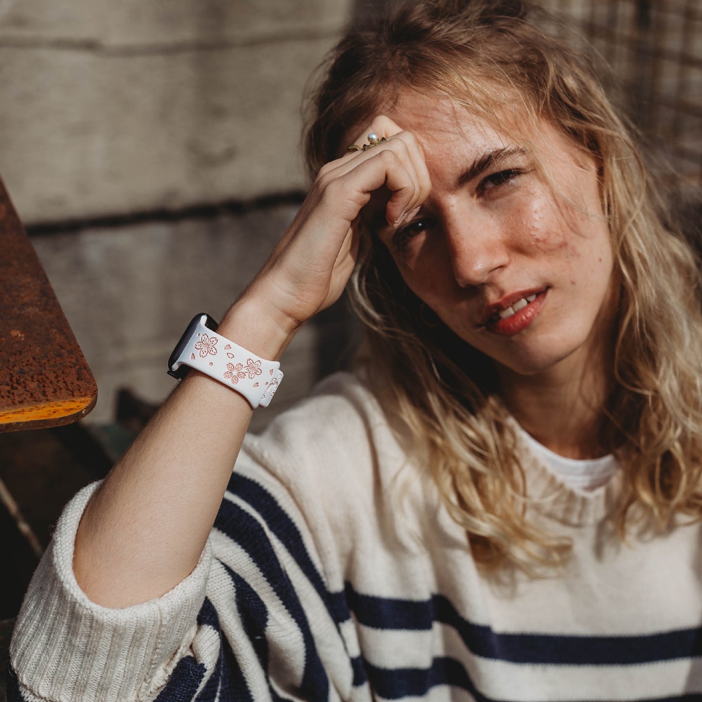 Eine Person mit langen blonden Haaren, die einen weiß-marineblauen gestreiften Pullover und ein Arktisband-Apple-Watch-Armband „Flores“ von Arktisband trägt, sitzt draußen, stützt den Kopf auf die Hand und blickt in die Kamera.