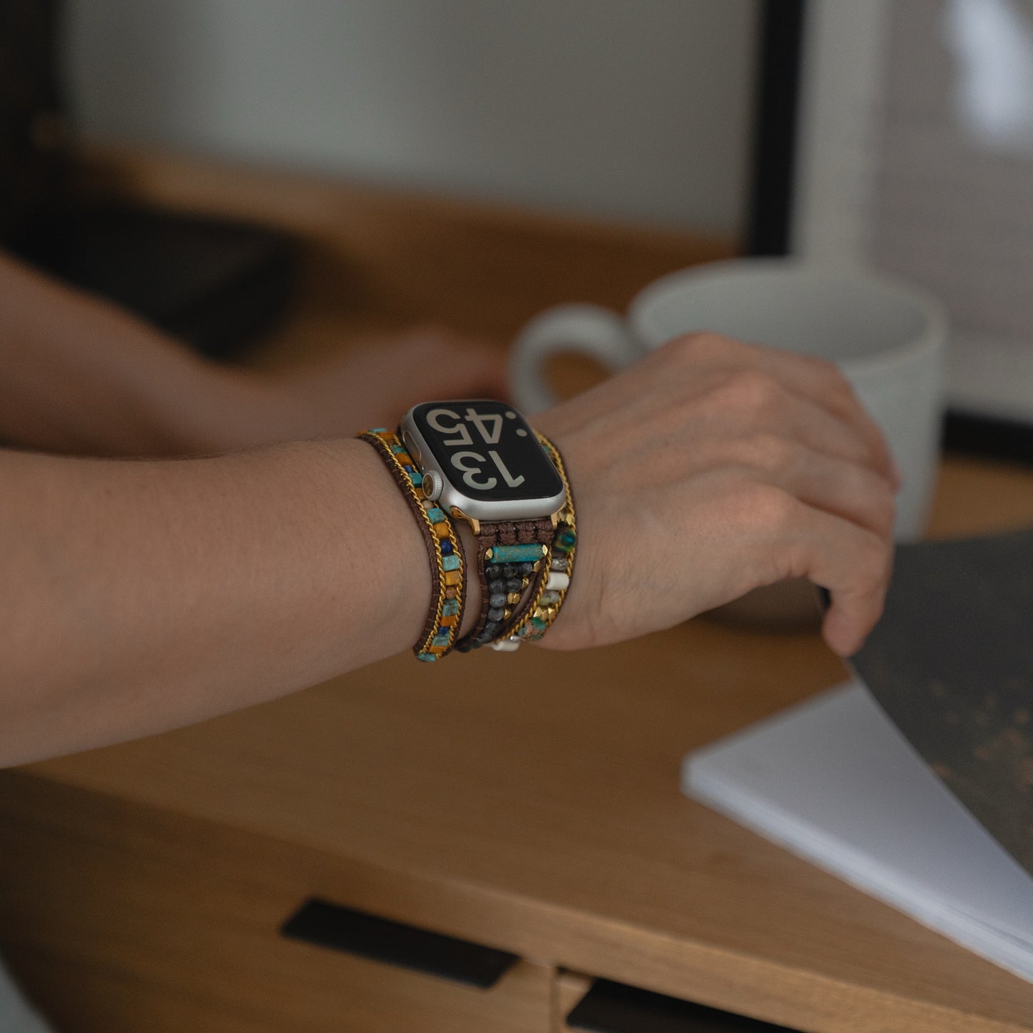 Eine Person trägt das arktisband Apple Watch Armband "Dream Catcher" und ein Boho-Armband, während sie nach einer weißen Tasse auf einem Holztisch greift und so mühelos intelligente Technik mit stilvollem Design verbindet.