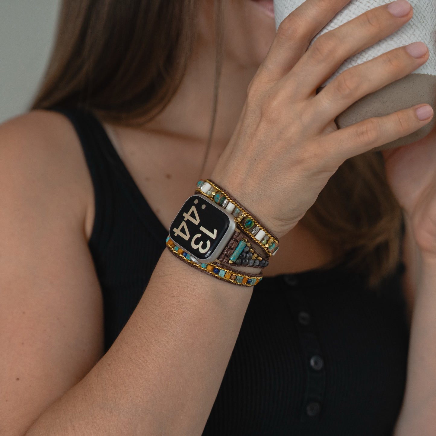 Eine Frau in einem schwarzen Tank-Top trinkt aus einem Becher und trägt dazu das arktisband Apple Watch Armband "Dream Catcher" und von der Boho-Kultur inspirierte Perlenarmbänder von arktisband für einen eleganten Look.