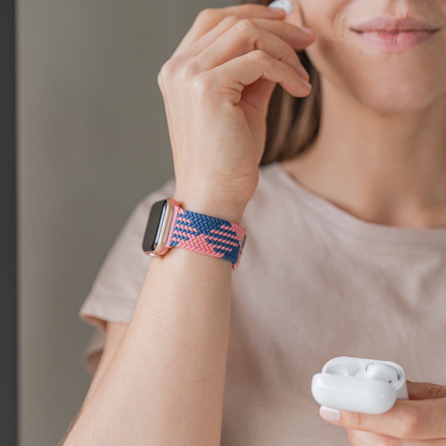 Eine Frau trägt ein beiges T-Shirt und eine Arktisband-Apple Watch mit dem geflochtenen Flex Loop TwoTone Armband. In einer Hand hält sie ein kabelloses Ohrhörer-Etui, während sie mit der anderen einen Ohrhörer in ihrem Ohr berührt.